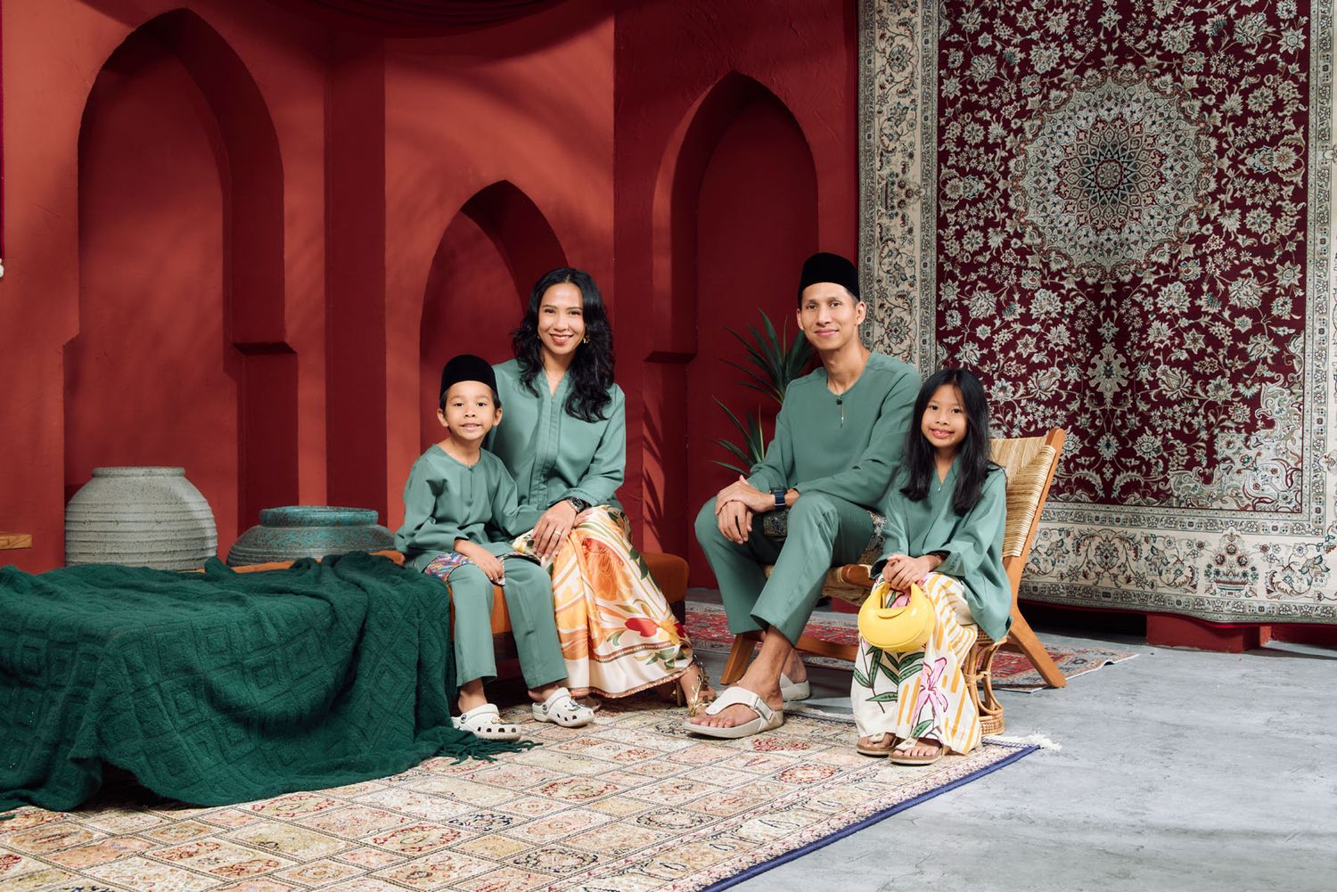 Family of four in matching green Baju Melayu and Baju Kurung seated in a red Moroccan-inspired photo set.