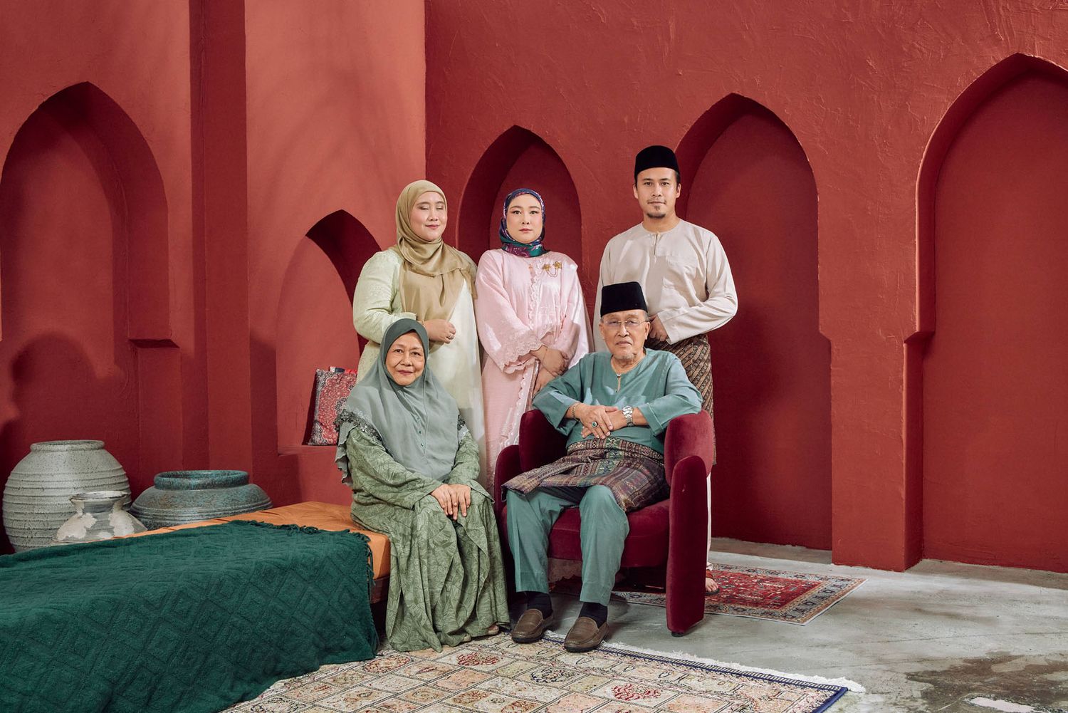 Multi-generational Malaysian family of five posing in traditional Raya outfits with red arched backdrop.
