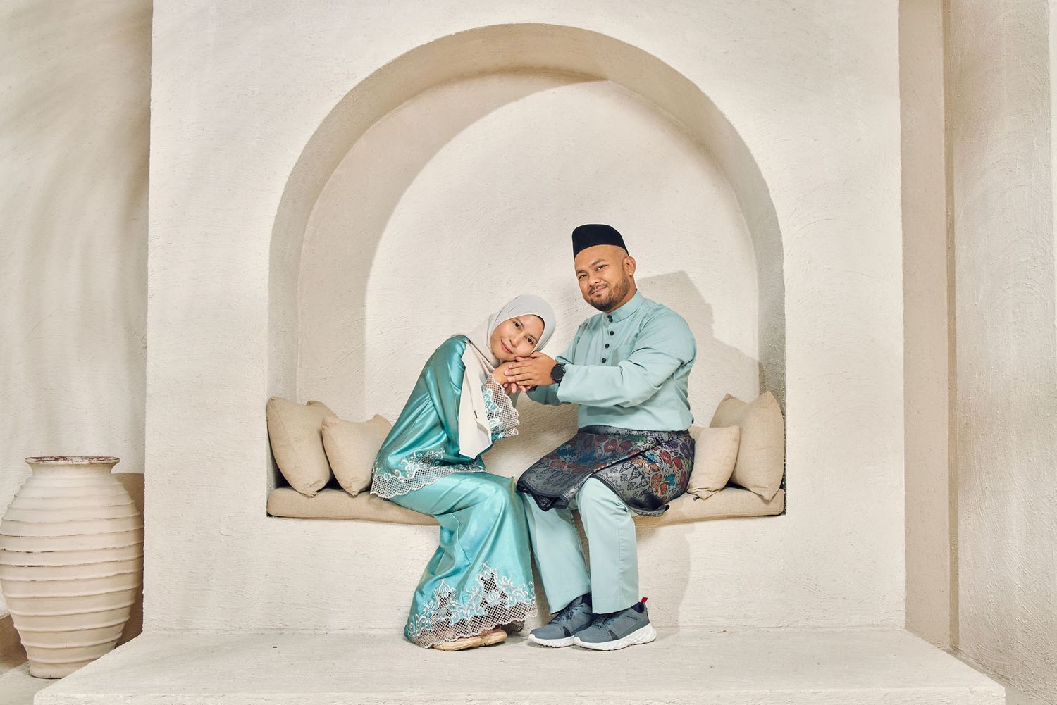 Couple in matching light blue traditional Malay baju kurung and baju melayu sitting in white arched alcove.