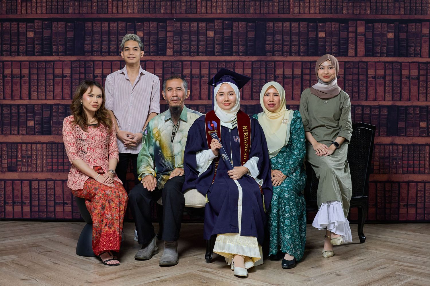 Family portrait with graduate in white gown and cap posed before bookshelf backdrop.