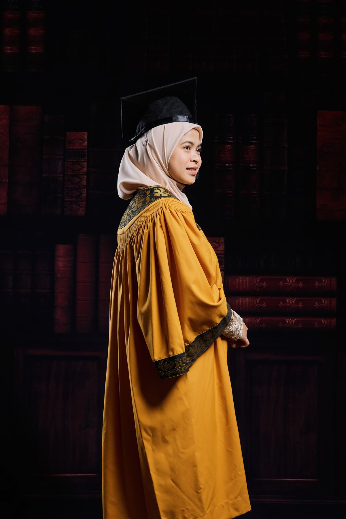 Muslim woman in golden graduation gown and hijab poses elegantly against a dark bookshelf backdrop.