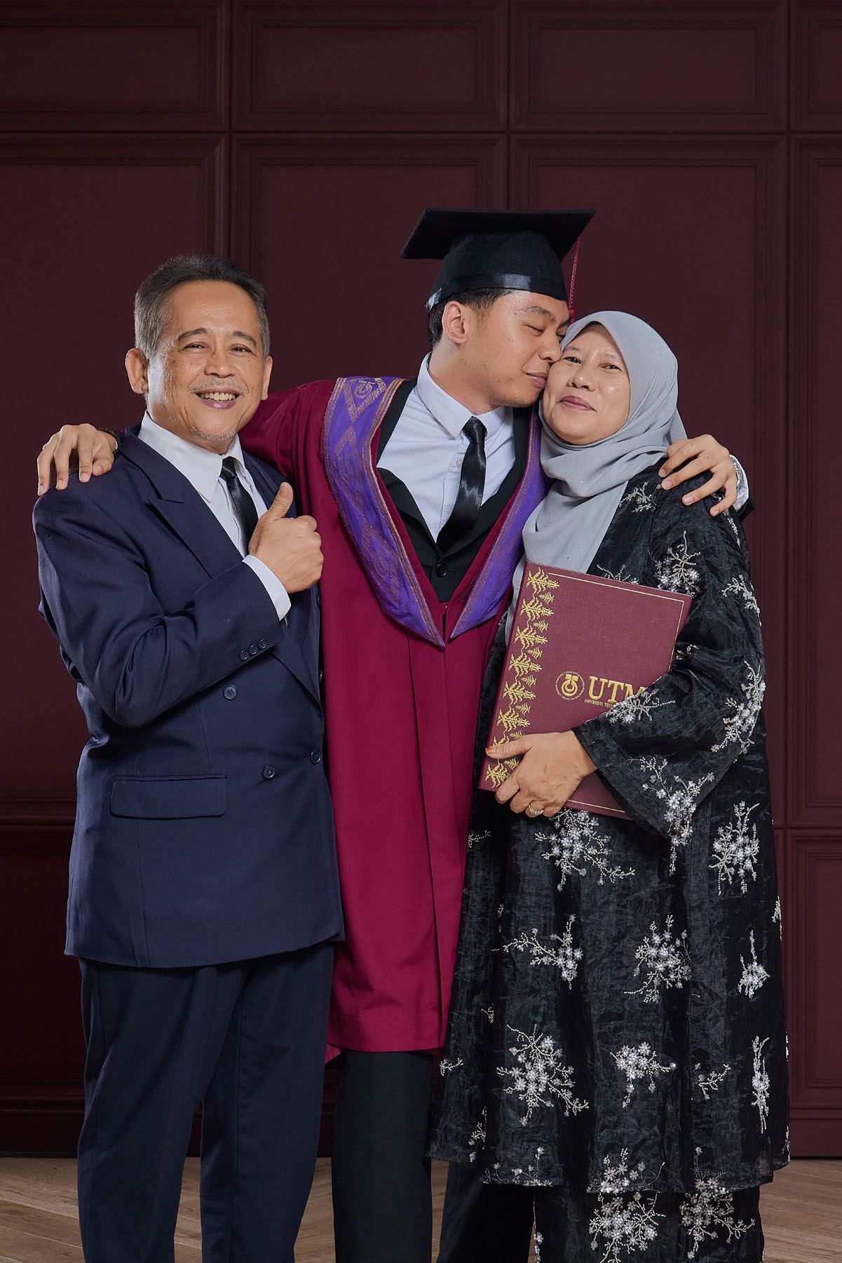 Graduate in maroon robe kisses mother while father gives thumbs up holding UTM diploma certificate.