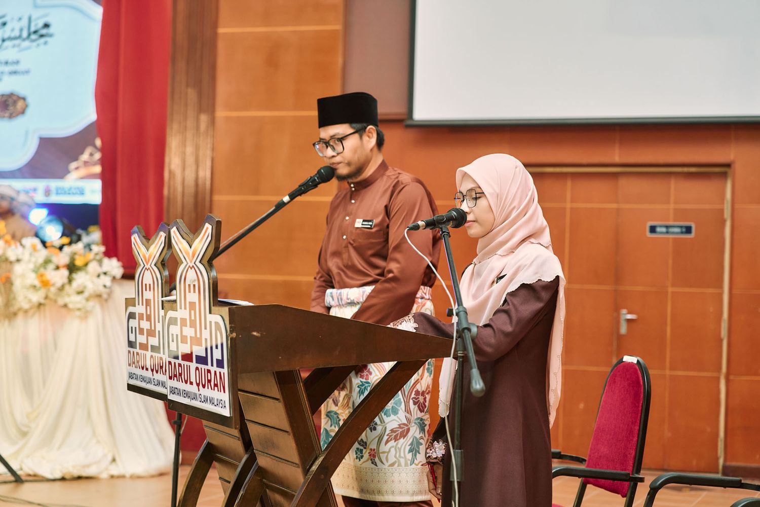A man and woman in traditional Malaysian attire speaking at a podium during a formal event.