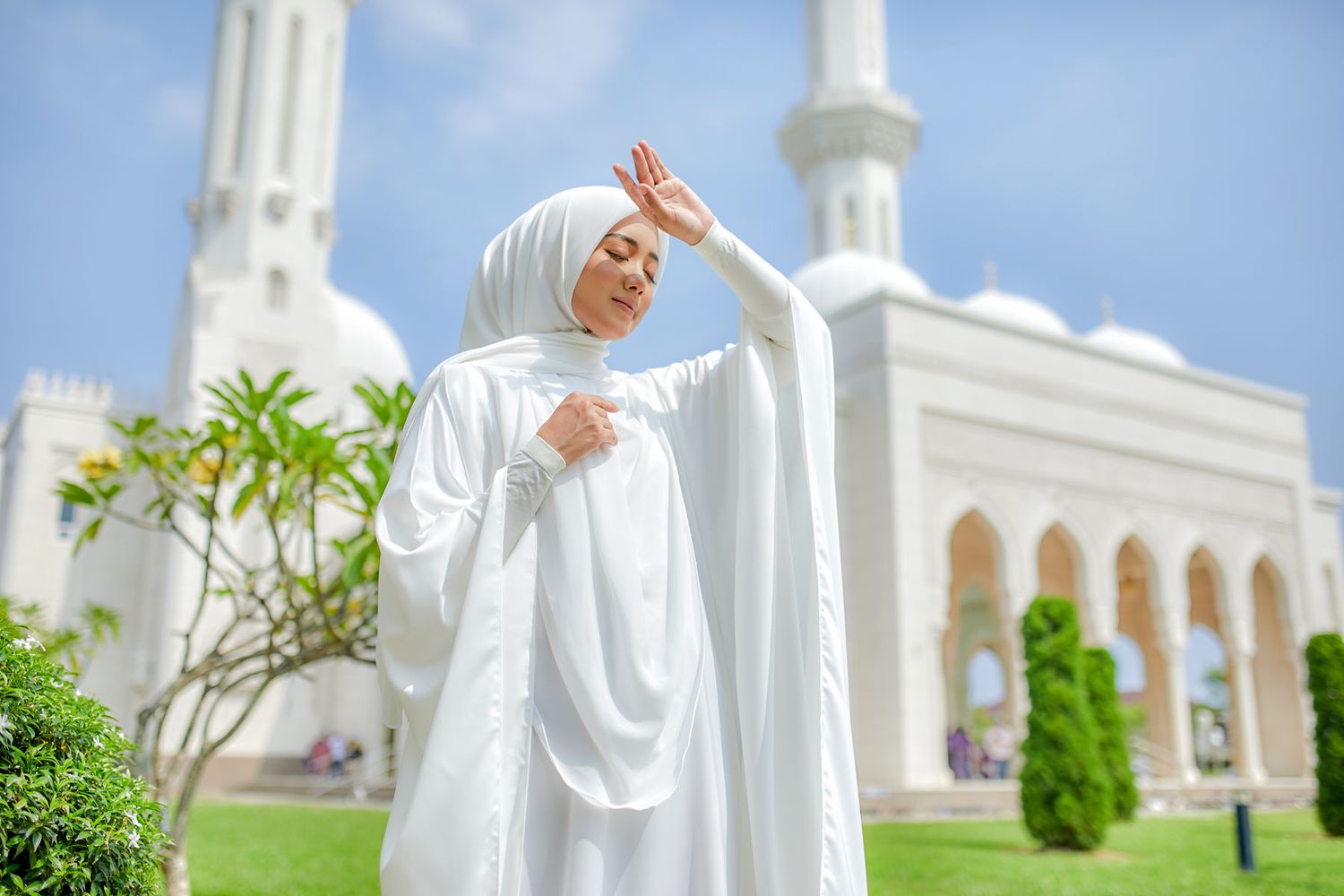 Muslim woman in white hijab and abaya posing gracefully in front of a grand white mosque.