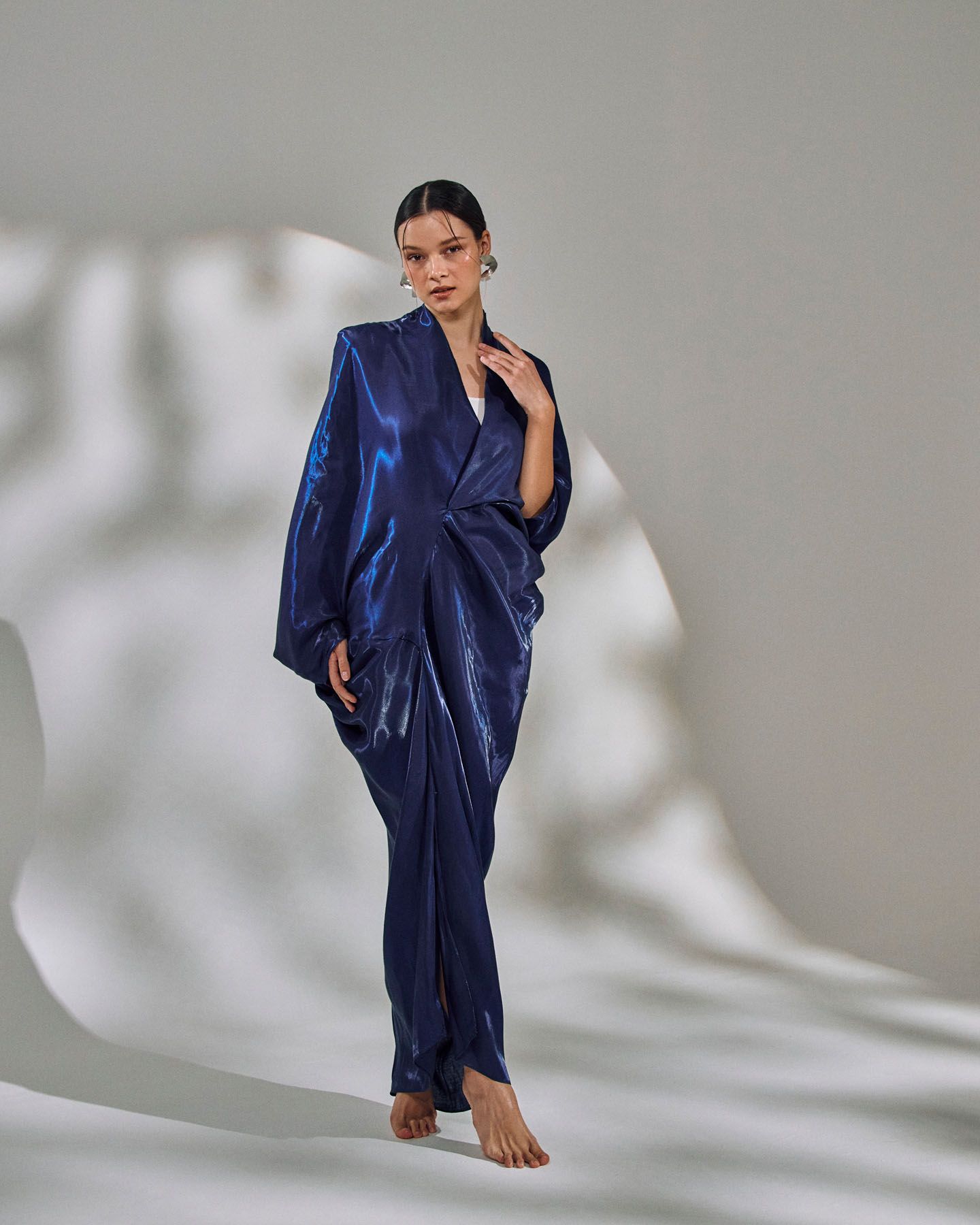 Model in a flowing navy blue metallic satin maxi dress with wide sleeves poses barefoot on gray backdrop.