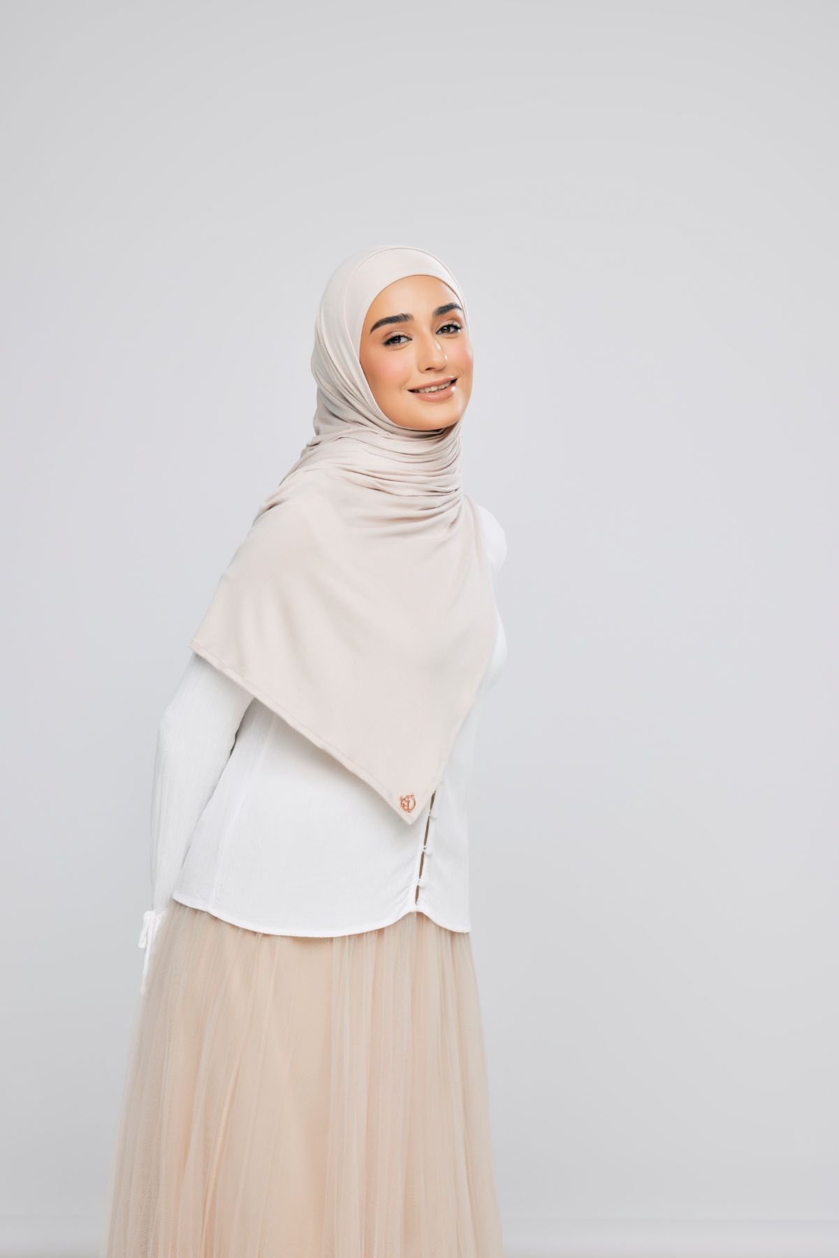 Young woman wearing a beige hijab, white top, and tulle skirt posing on light background.