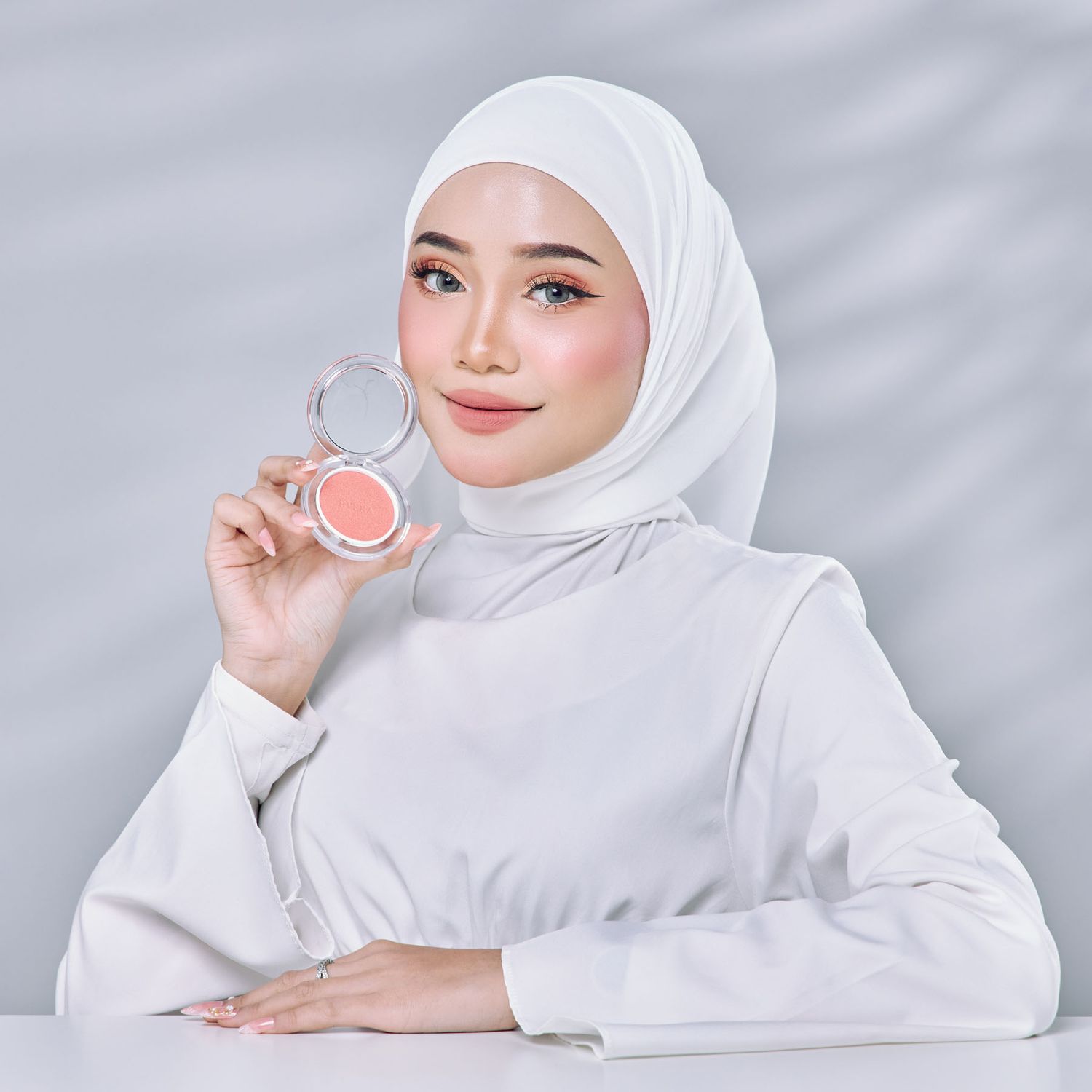 Woman in white hijab holding open compact blush powder product in bright studio setting.