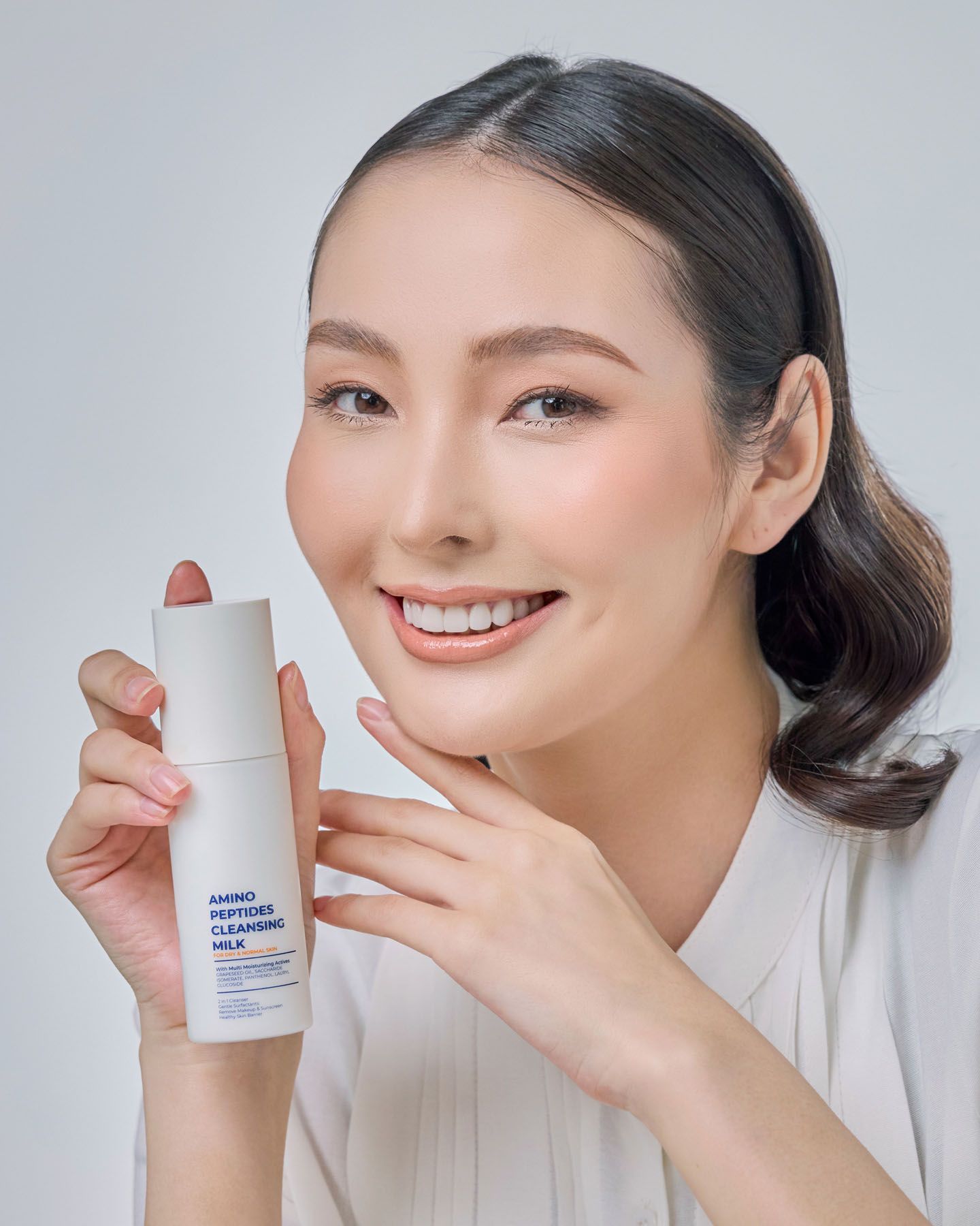 Smiling woman holding Amino Peptides Cleansing Milk with natural glowing makeup look.