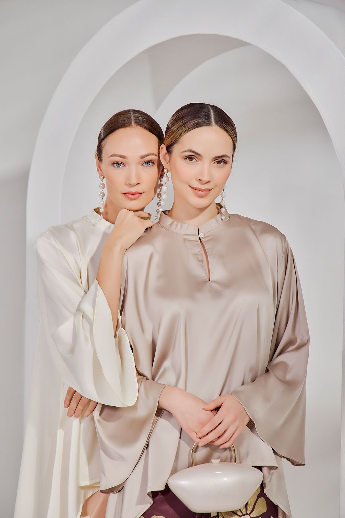 Two women in ivory and champagne satin kaftan tops pose elegantly in a white arched studio.