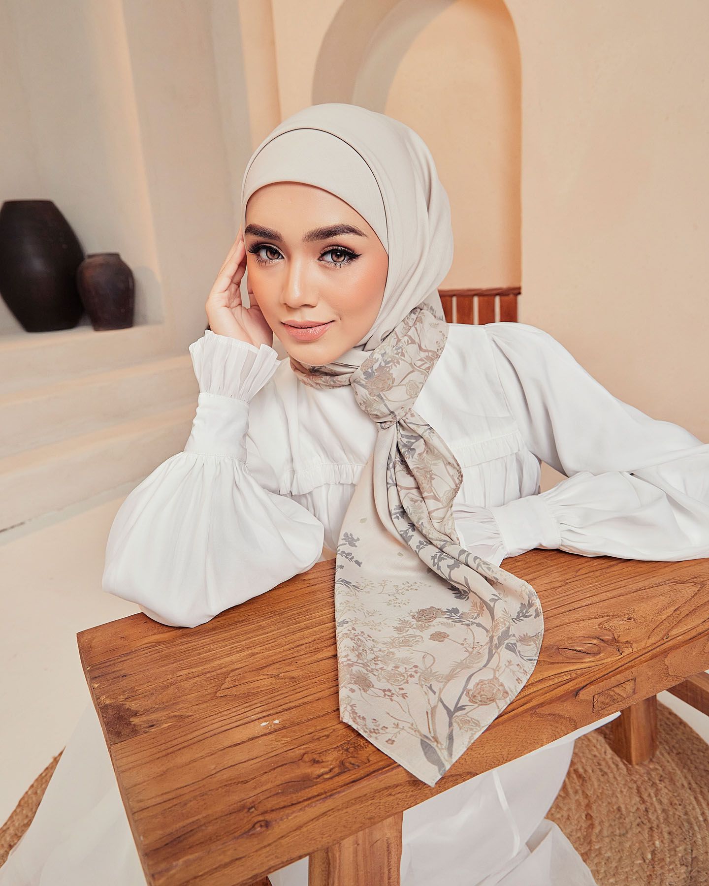 A woman in a beige hijab and white dress wears a soft floral printed scarf while resting at a wooden table.
