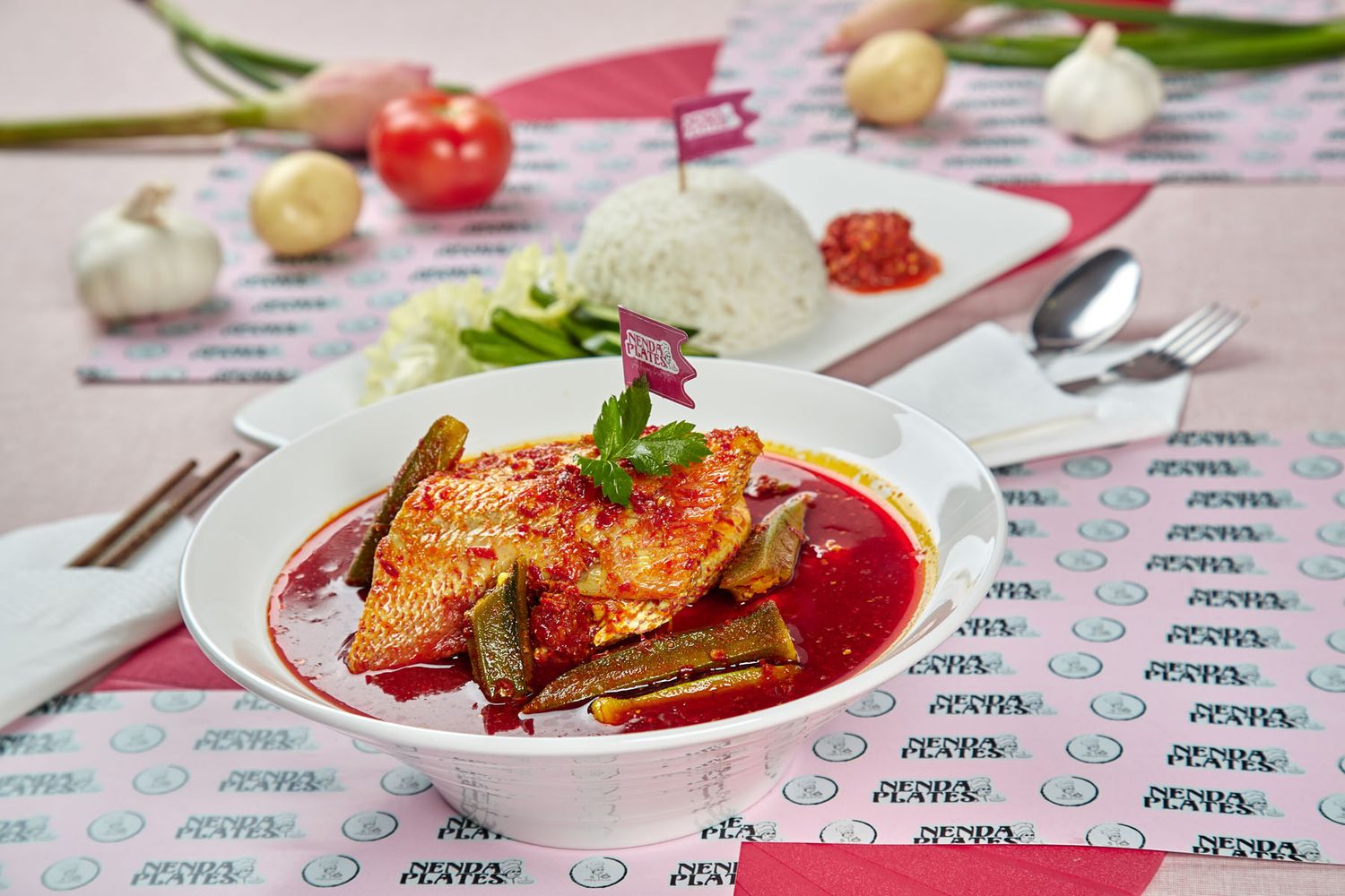 Malaysian fish curry in red sauce served in white bowl with steamed rice and sambal on patterned cloth.