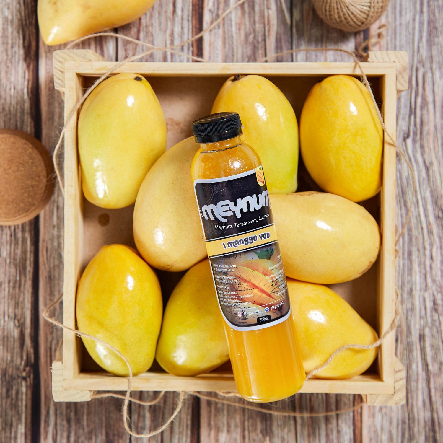 A bottle of Meynum mango juice nestled among fresh yellow mangoes in a rustic wooden crate.