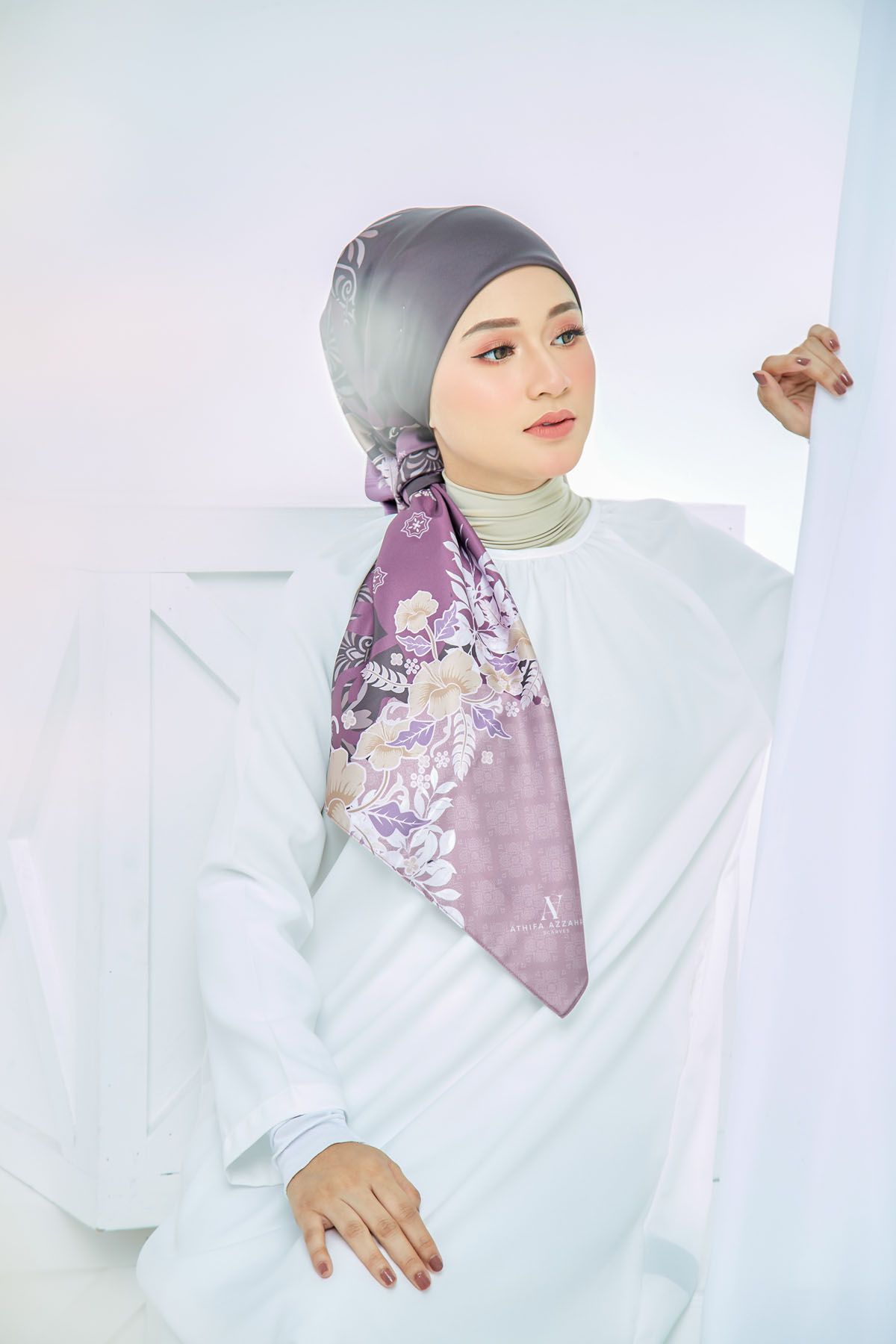 Woman in white outfit wearing gray hijab with purple floral silk scarf in elegant modest fashion pose.