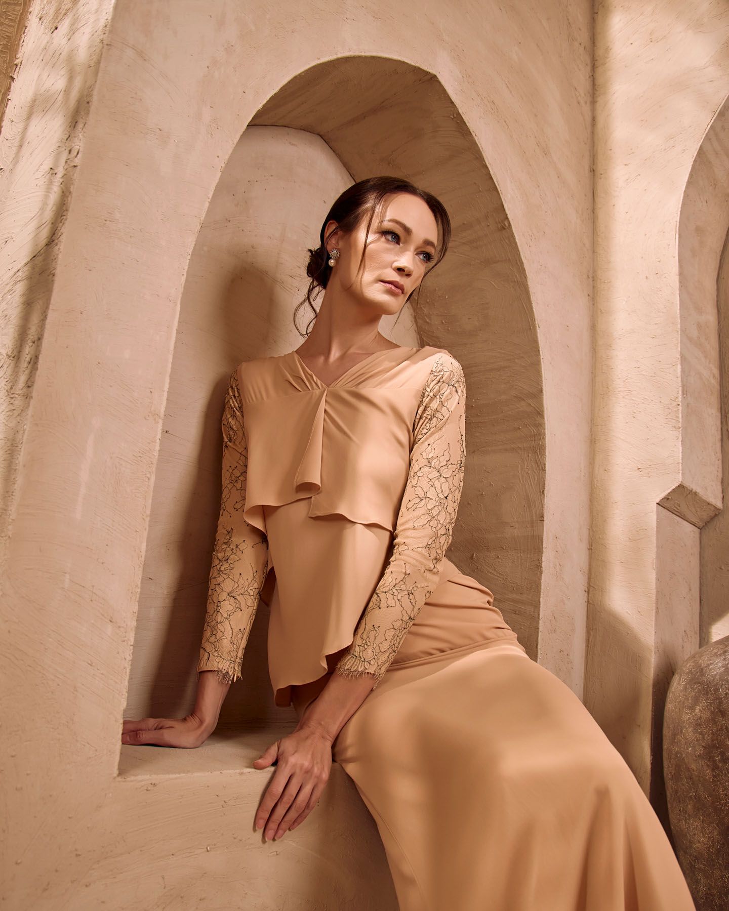 Woman in champagne gold lace-sleeve peplum dress sits in layered stone archway niche, elegant pose.