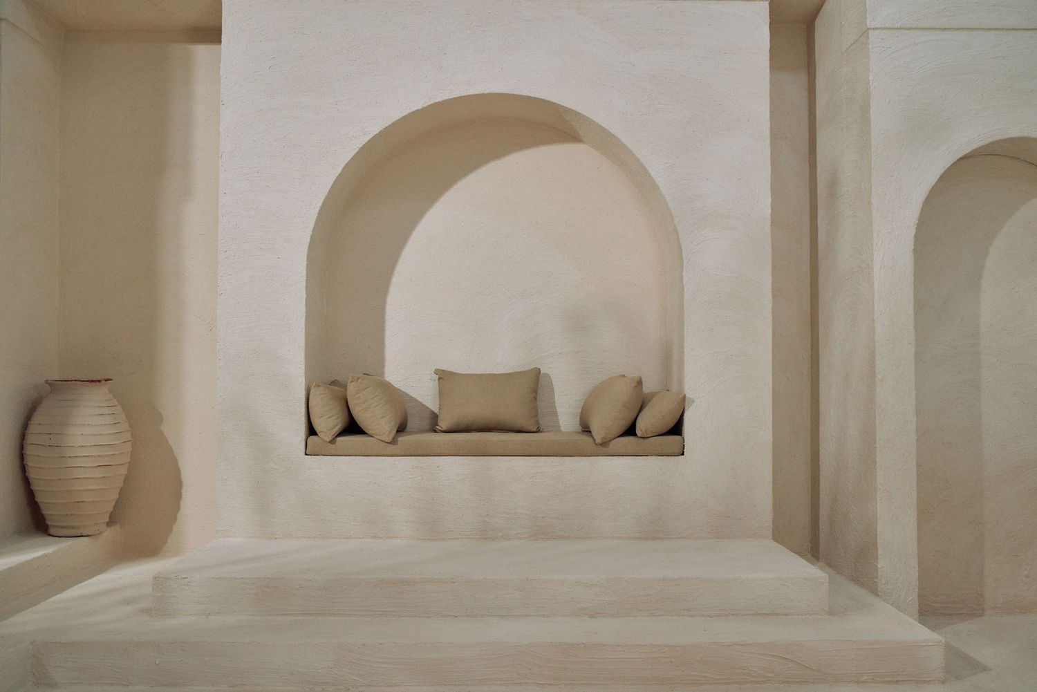 Serene minimalist interior with beige plaster arched niches, linen cushions, and rustic ceramic vases.