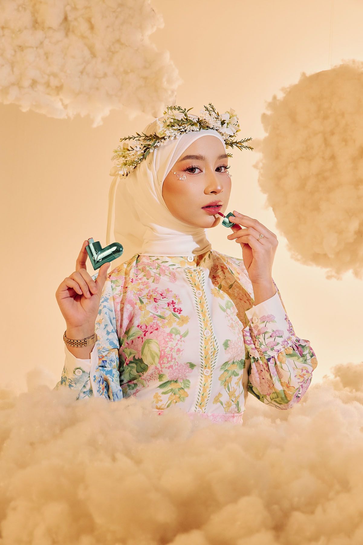 Muslim woman in white hijab with floral crown holds Ayla Cosmetics Twist lip gloss among clouds.