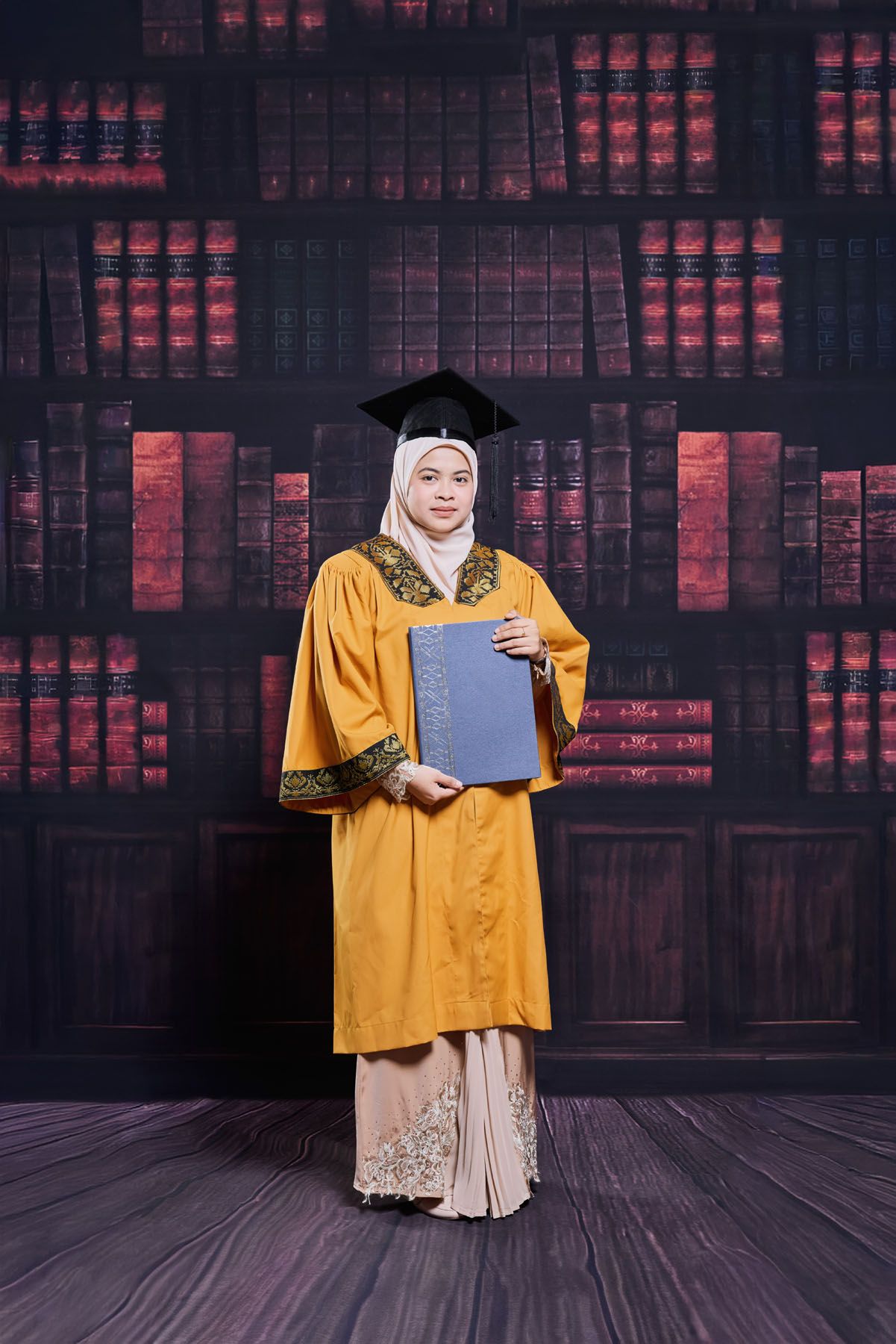 Muslim woman graduate in golden robe and hijab holding diploma book against library bookshelf backdrop.