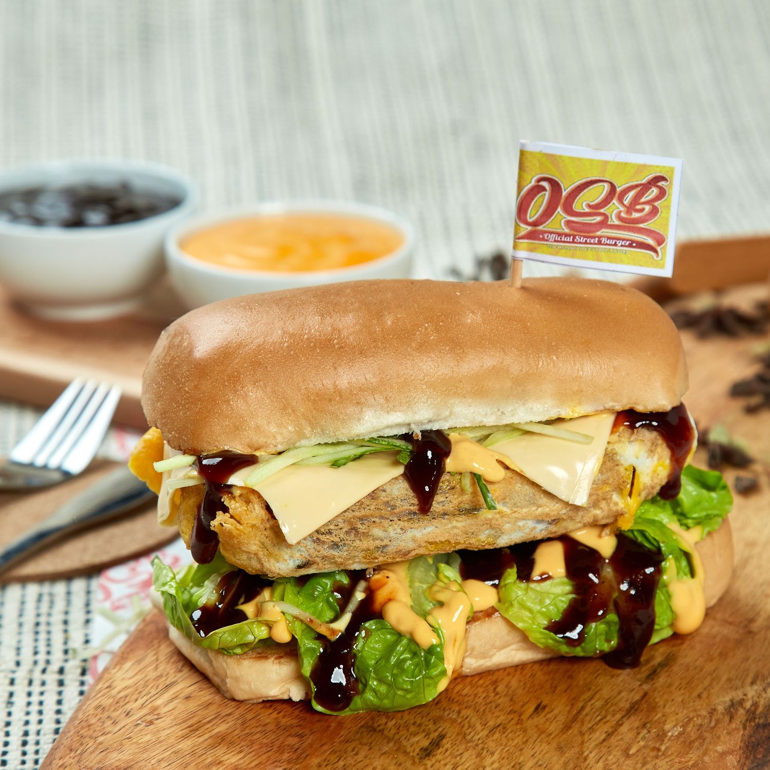 OCS branded gourmet burger with melted cheese, lettuce, and BBQ sauce on wooden board with dipping sauces.