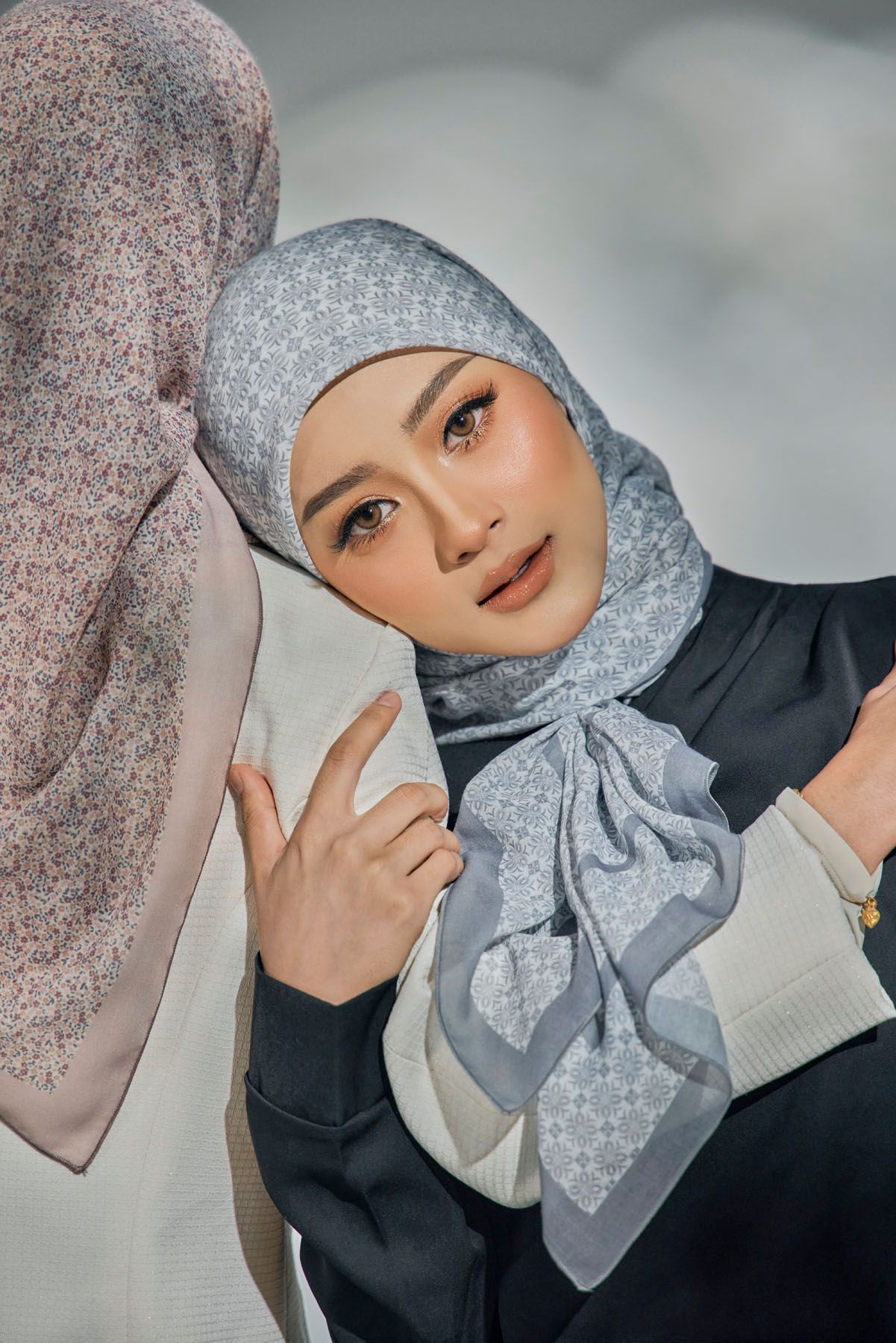 Close-up of woman in blue patterned hijab with elegant makeup posing near another hijab model.