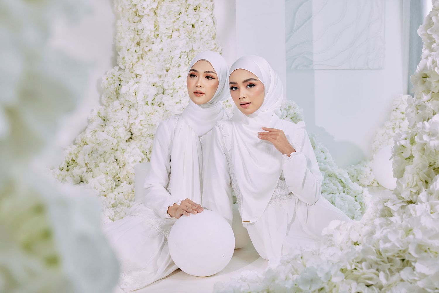 Two women in matching white hijabs and outfits seated among white floral decorations.
