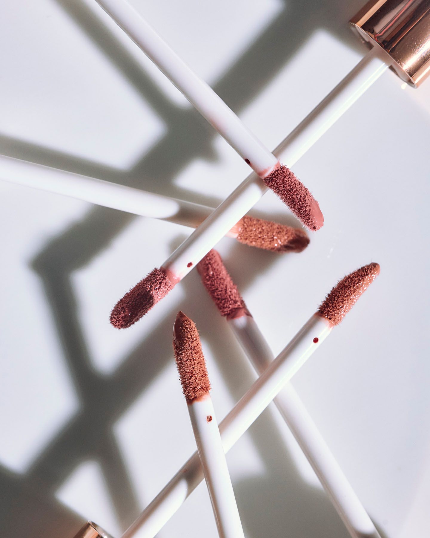 Close-up of multiple lipgloss wands with rose-toned product on white applicators casting geometric shadows.