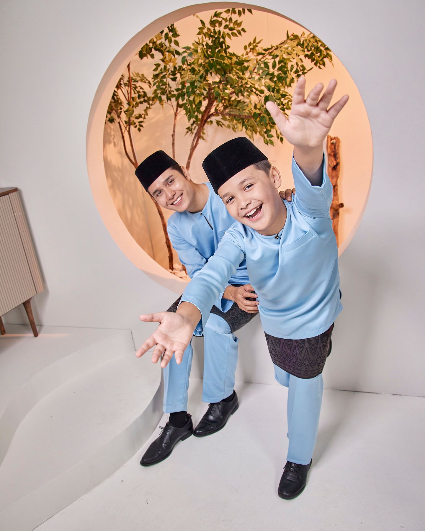 Two boys in matching light blue baju melayu wave playfully in a modern studio setting.