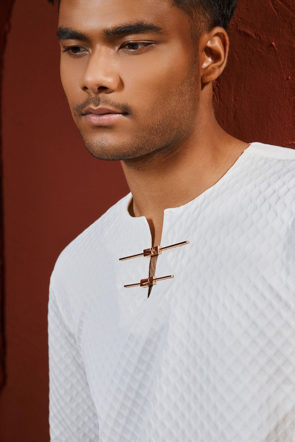Close-up of man in white quilted baju melayu with gold traditional clasp against a red wall.