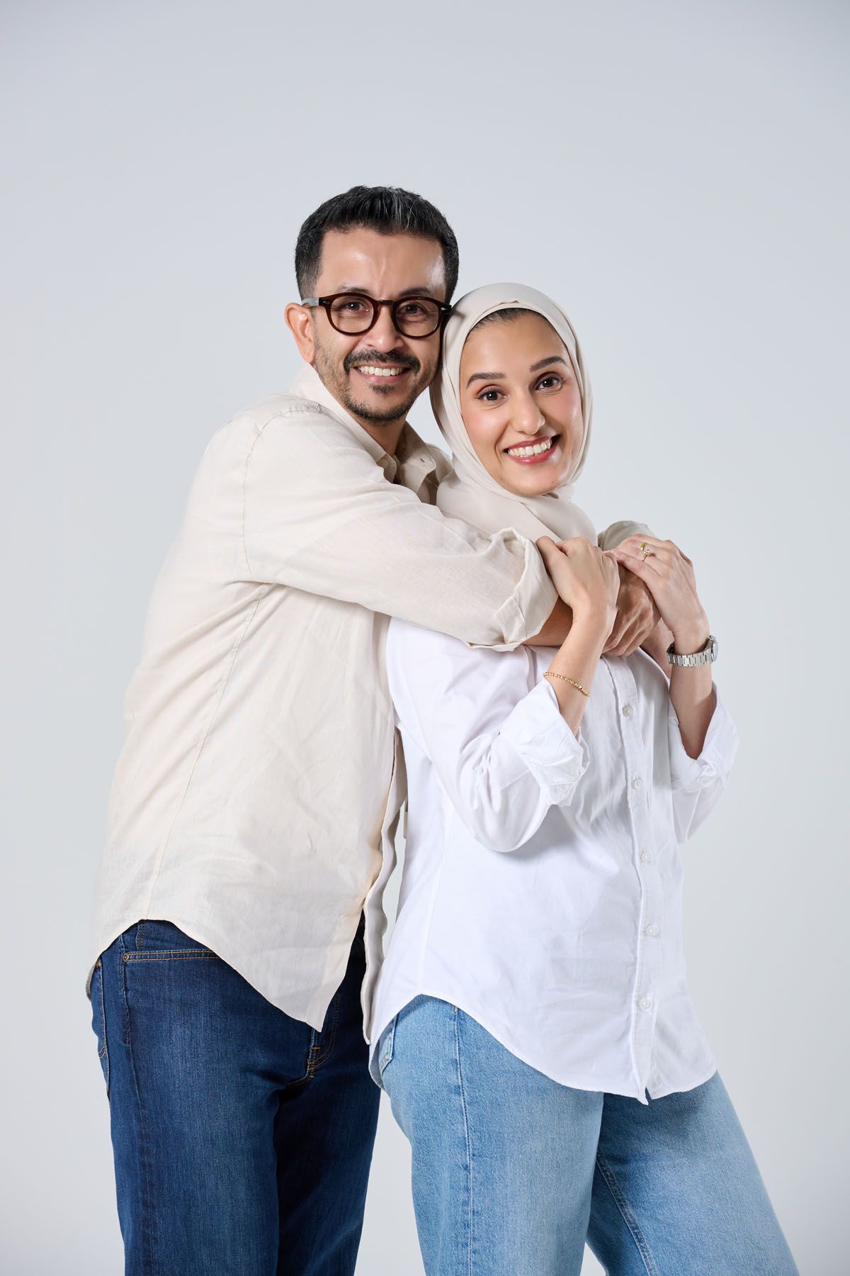 Smiling Muslim couple embracing, woman in hijab and white shirt, man in glasses and beige shirt, jeans, white background.