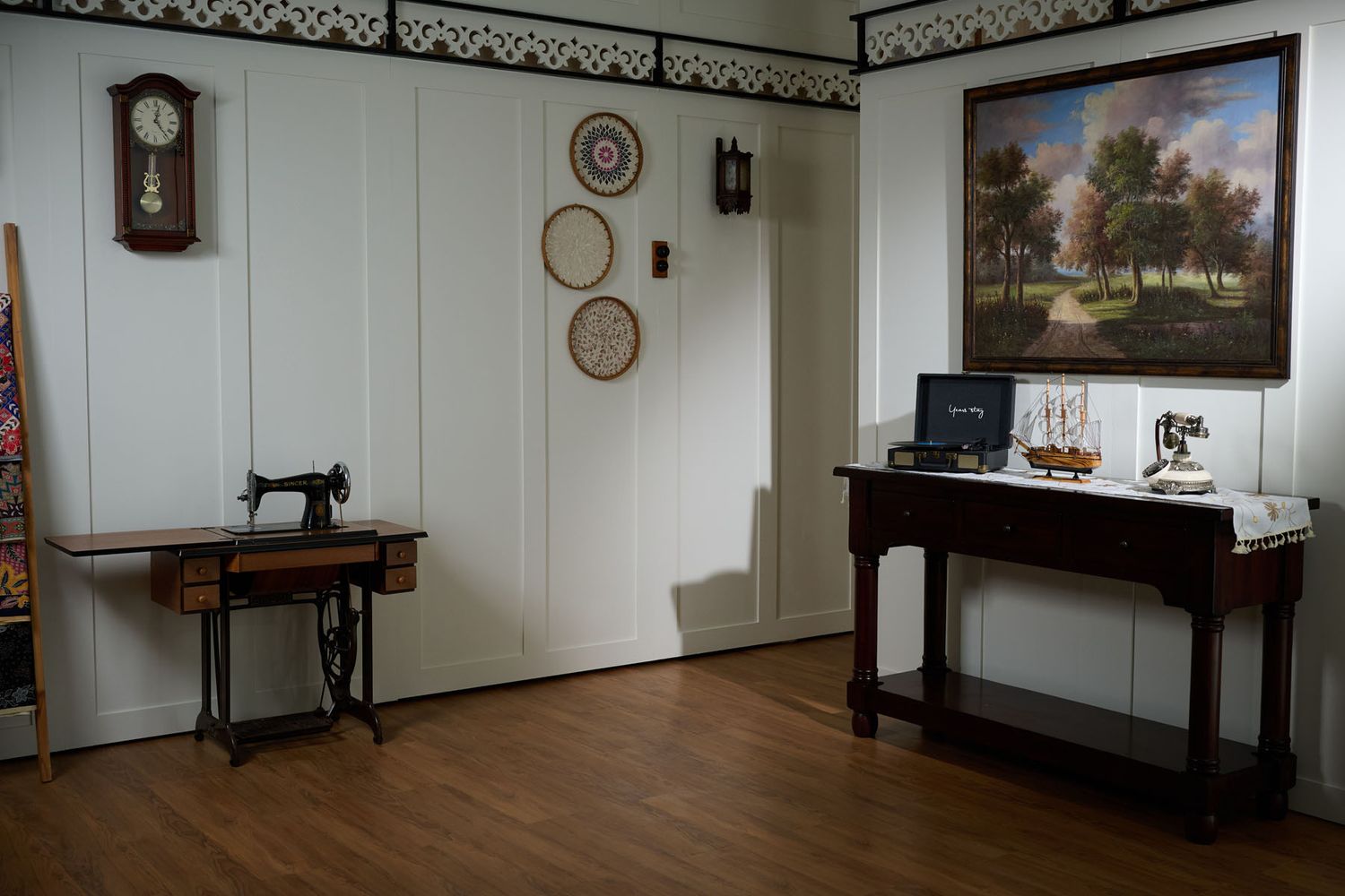 Vintage room display with antique sewing machine, wall clock, decorative plates, landscape painting, and wooden console table.