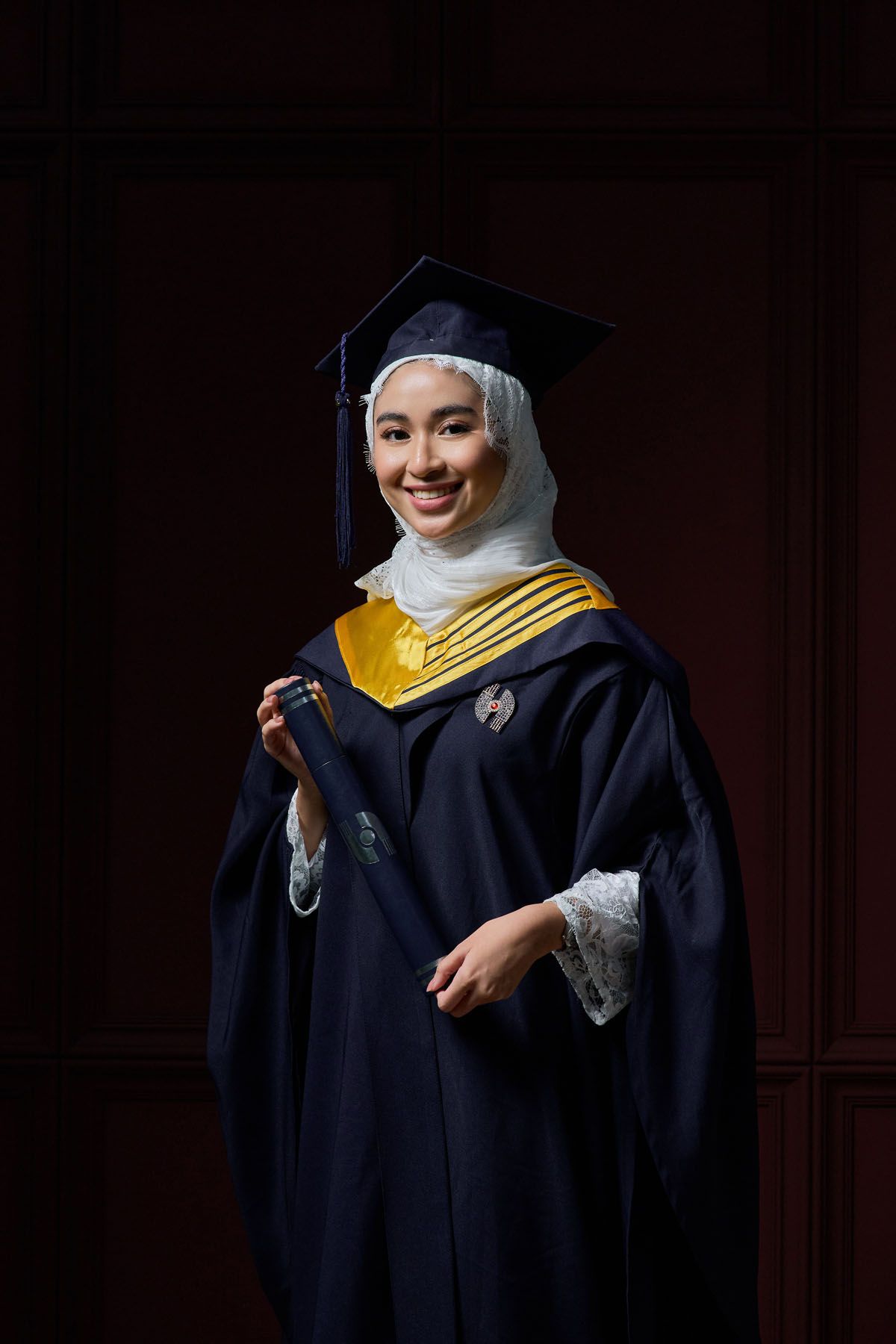 Smiling female Muslim graduate in black gown with yellow stole and white hijab holds diploma tube.