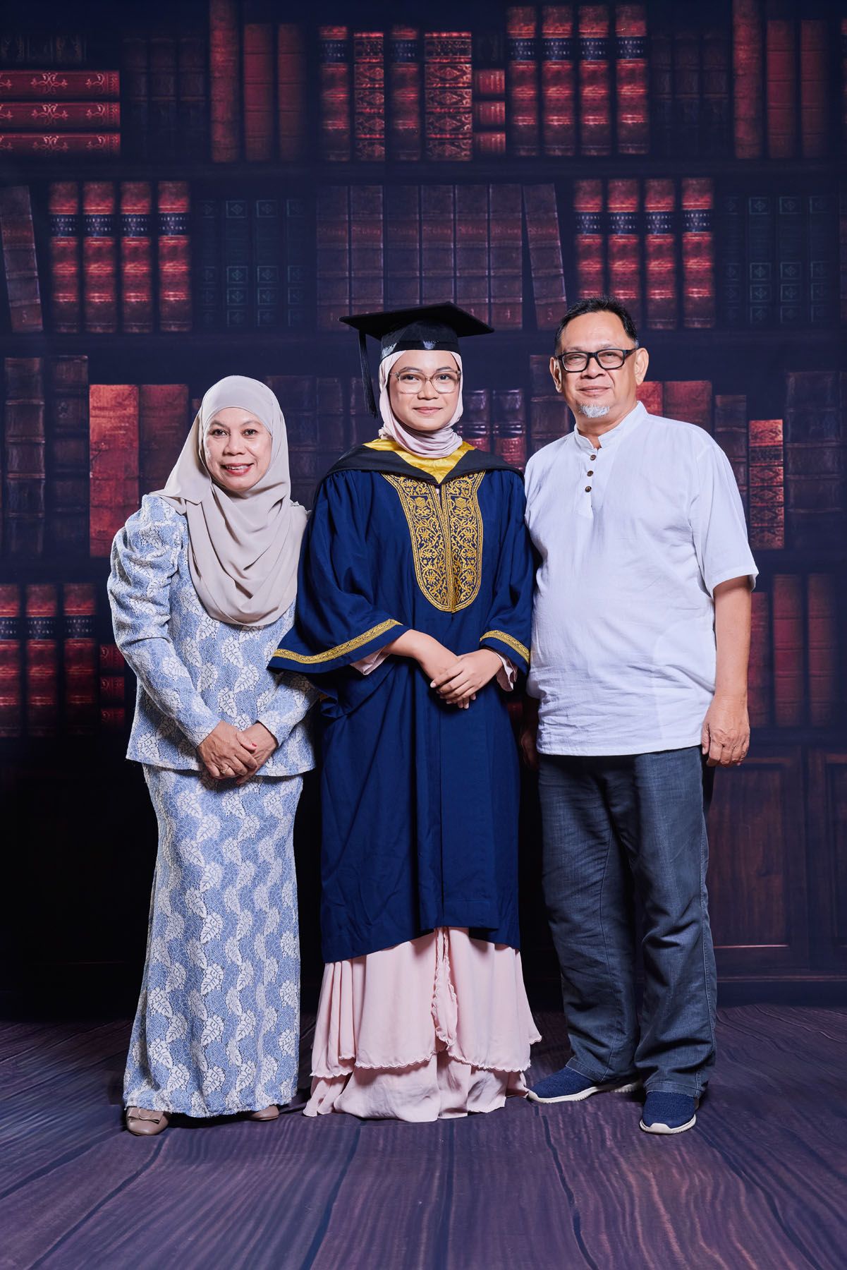 Female graduate in navy robe standing with parents before bookshelf backdrop.
