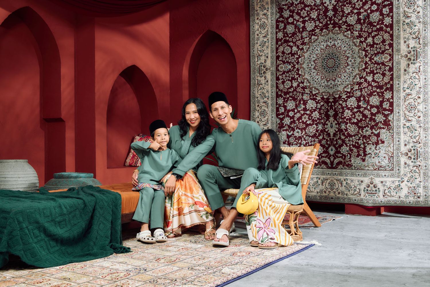 Family of four in matching green traditional Malay outfits posing casually in a red Moroccan-themed studio.