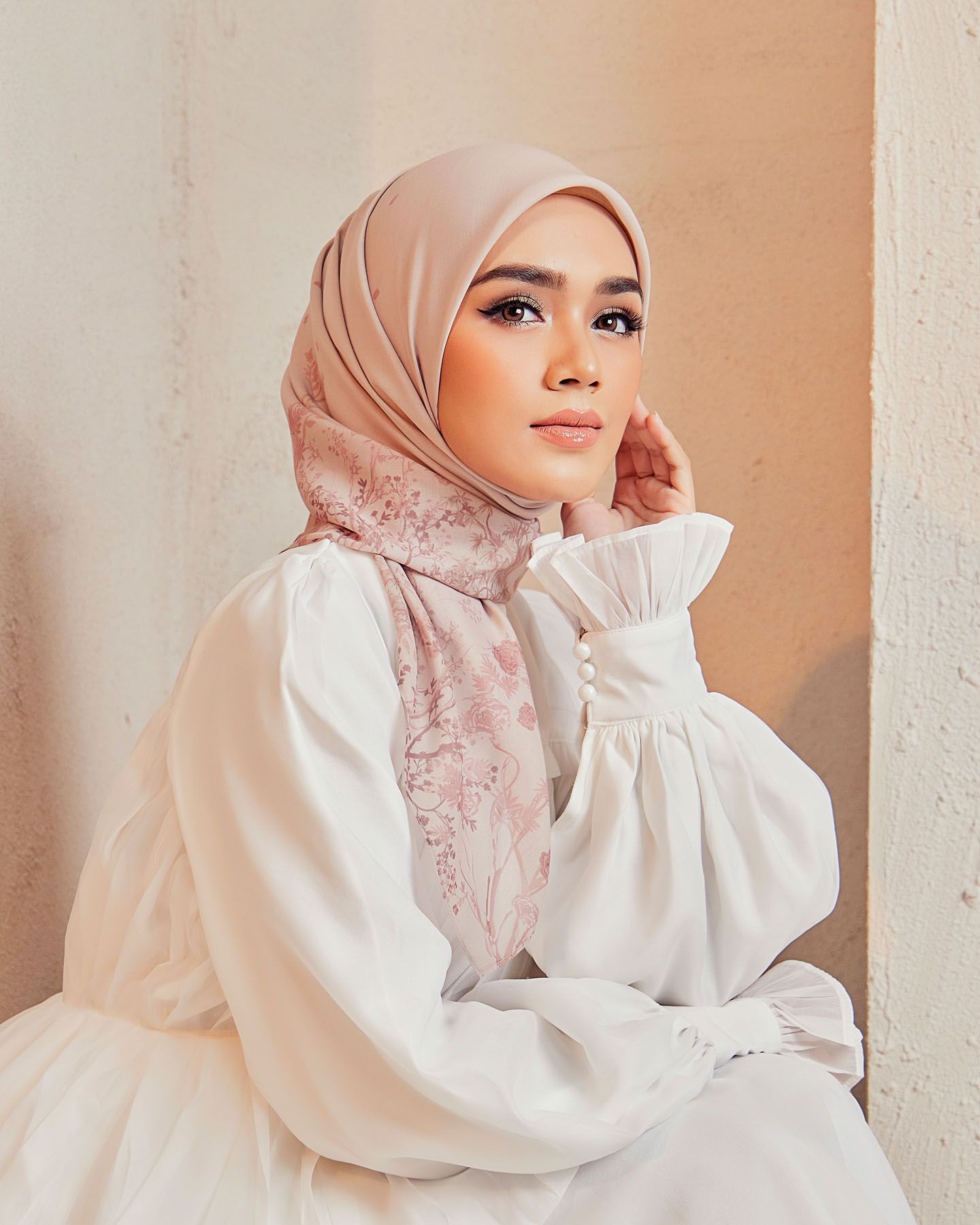 A woman in a blush pink hijab with floral lace detail and white dress poses gracefully in warm natural light.