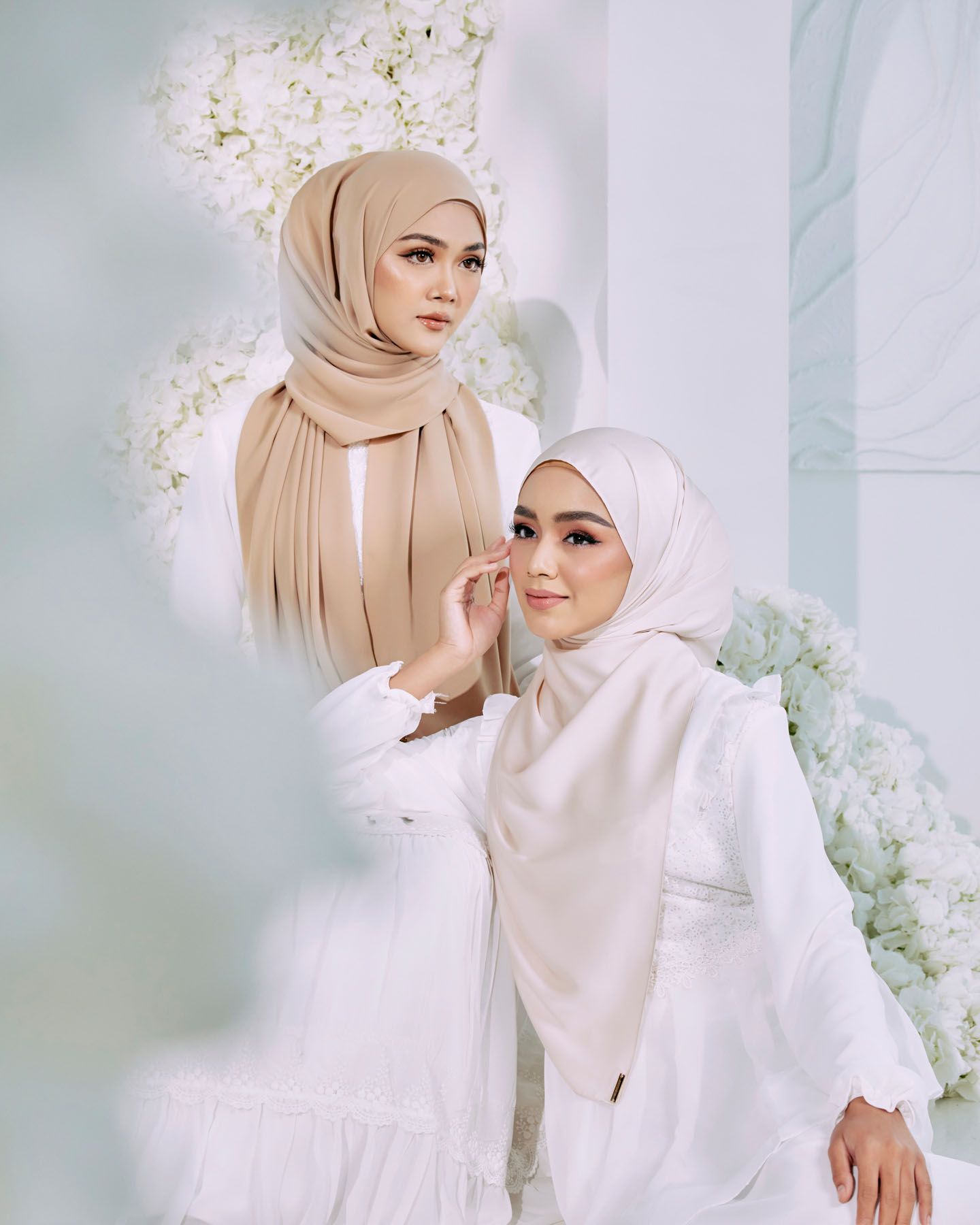 Two women in beige and white hijabs wearing white outfits posed before white floral backdrop.