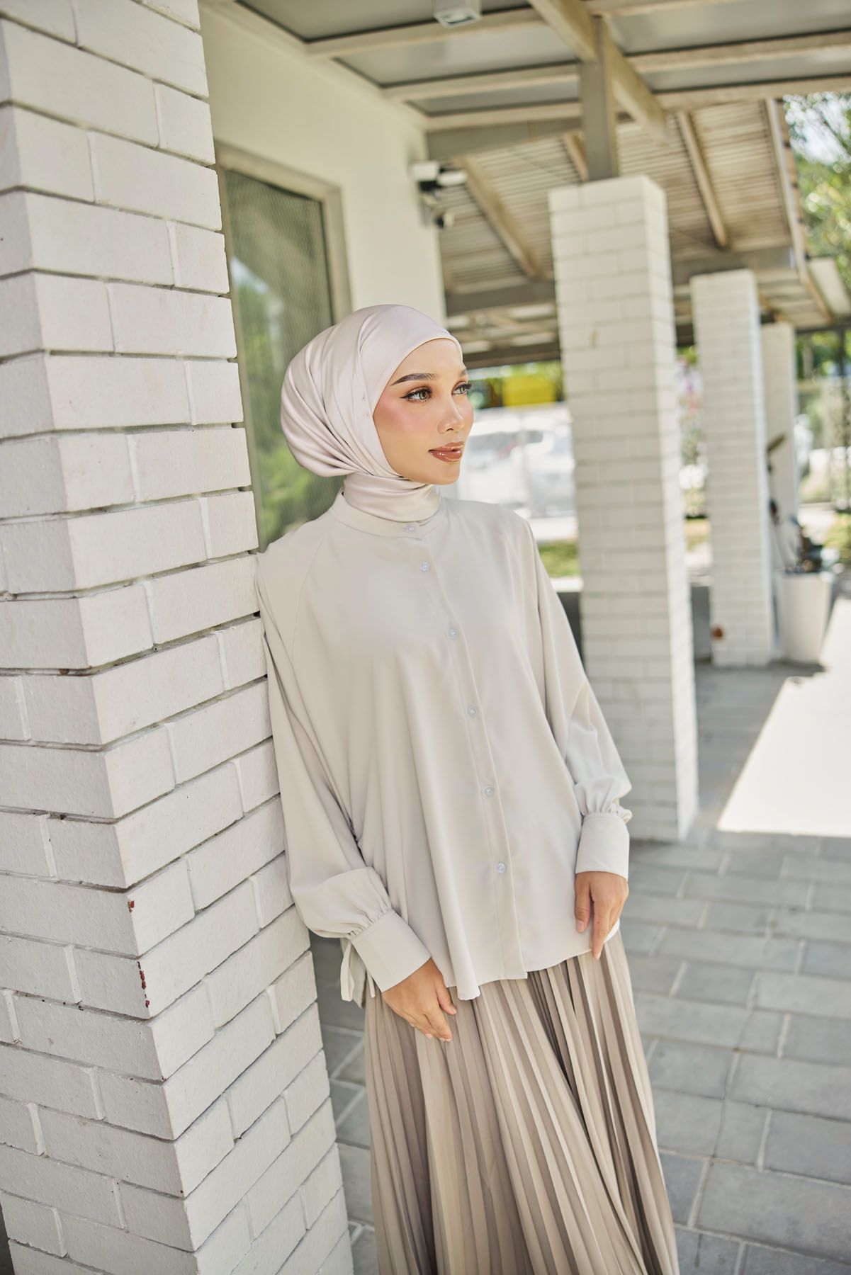 A woman in a white hijab and beige pleated skirt leans against a white brick wall outdoors.