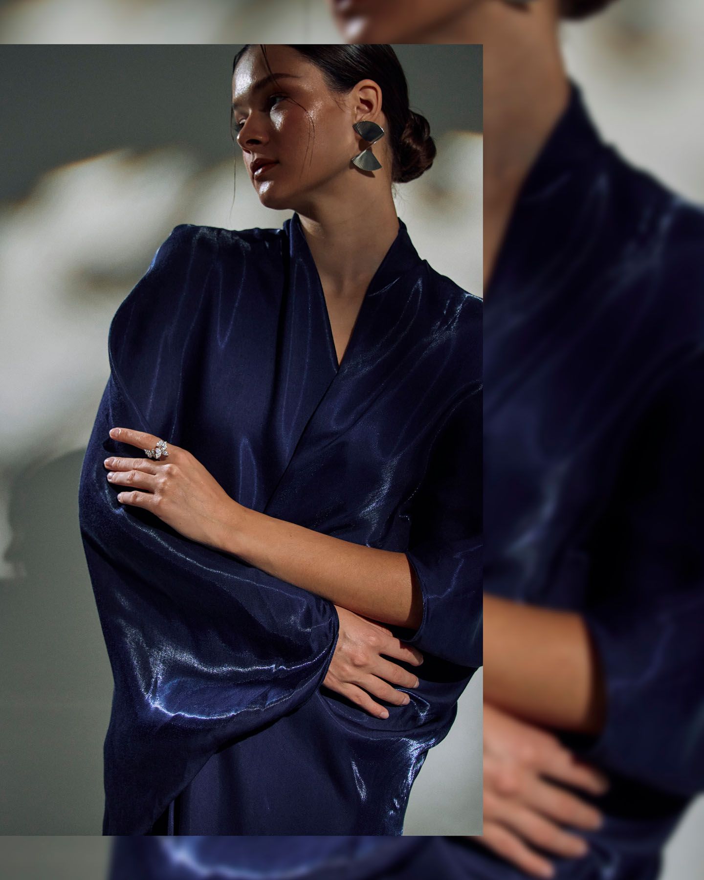 Close-up of woman in navy blue satin kimono with dark statement earrings and silver ring, arms crossed.