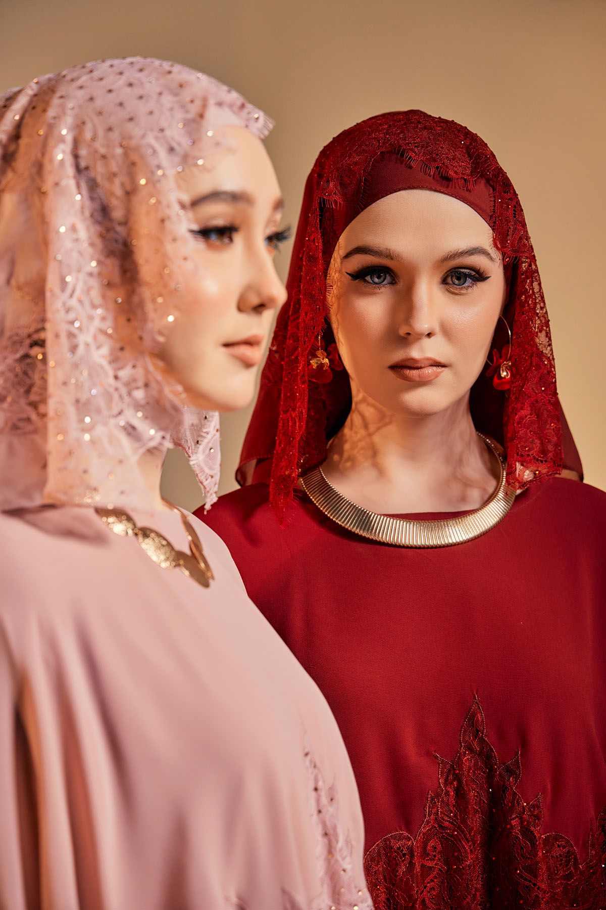 Two Muslim women in pink and red lace hijabs and elegant dresses posing closely together.