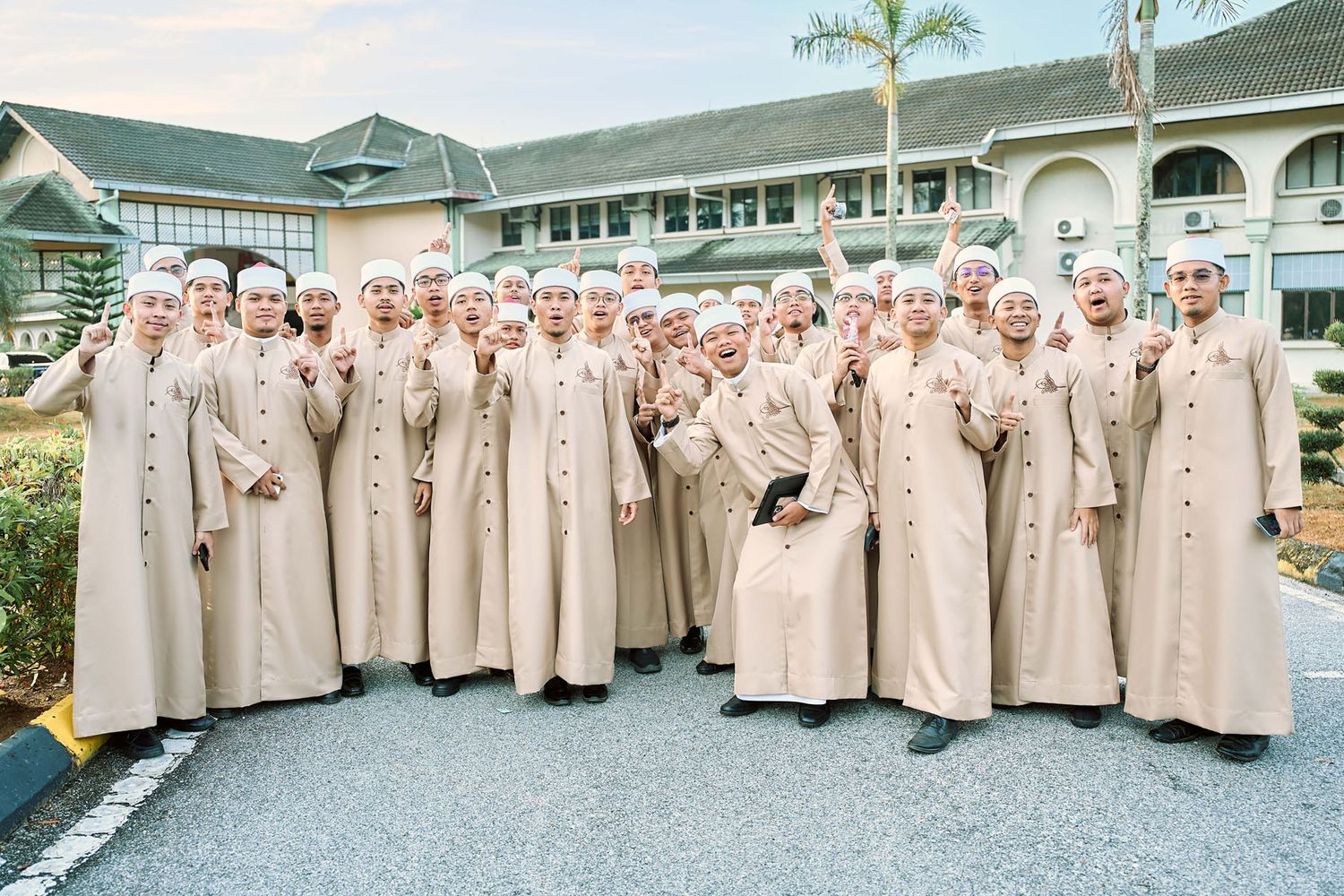 Large group of people in matching beige Islamic robes and white caps posing outdoors at a formal religious gathering.