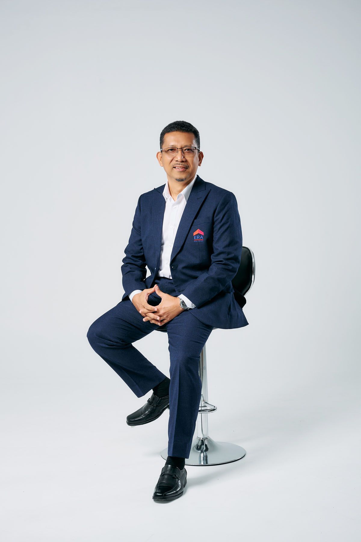 Professional man in navy blue suit sitting on bar stool, smiling confidently against white background.