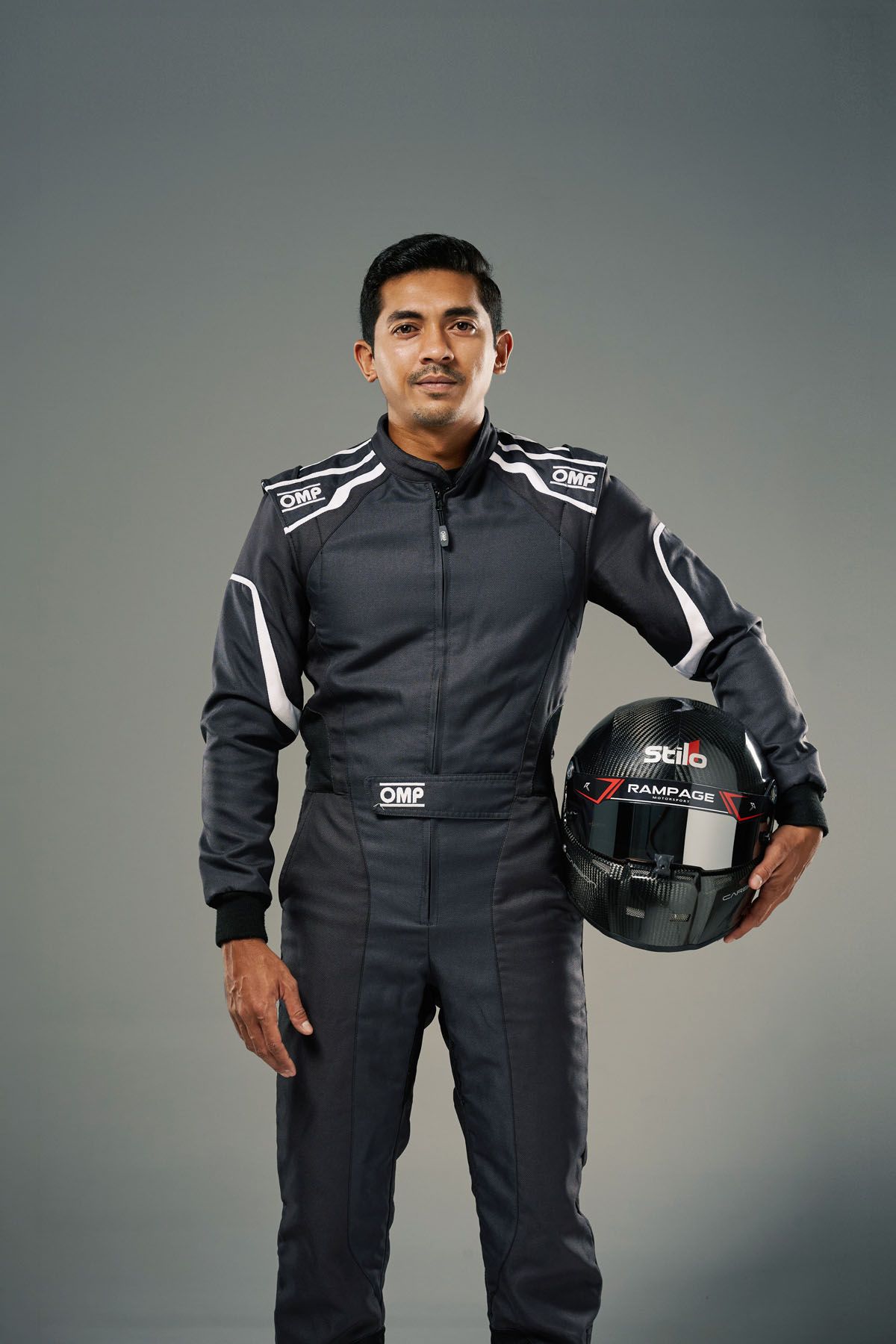 A racing driver in a black OMP suit holds a Stilo helmet against a gray studio background.