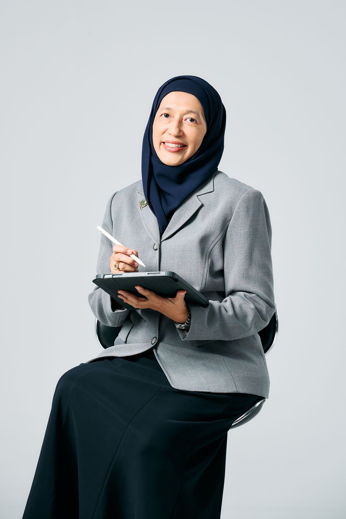 Smiling Muslim businesswoman in gray blazer and hijab sitting while writing notes in a planner.