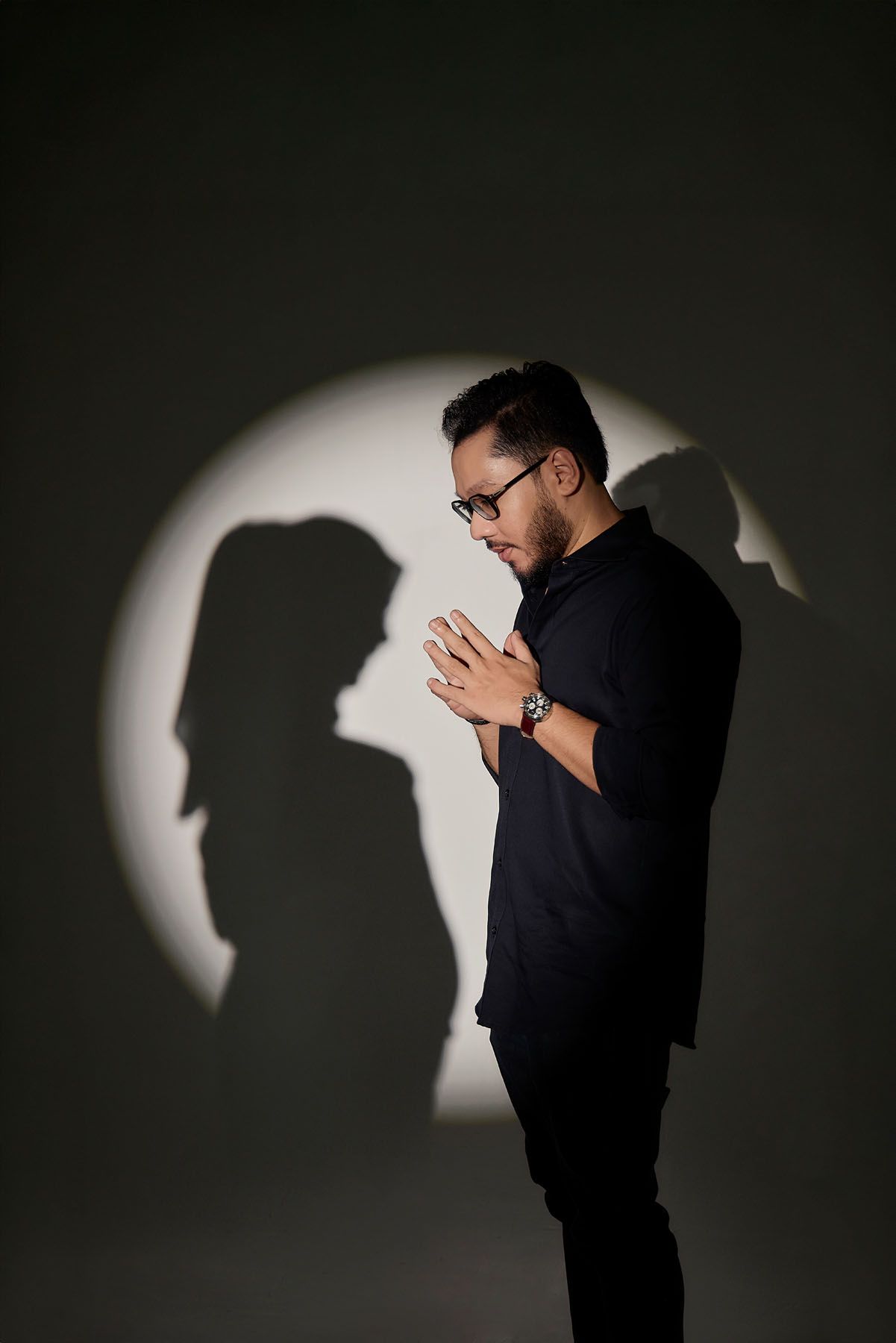 Man in black shirt praying with hands clasped, dramatic shadow silhouette of woman on grey wall.