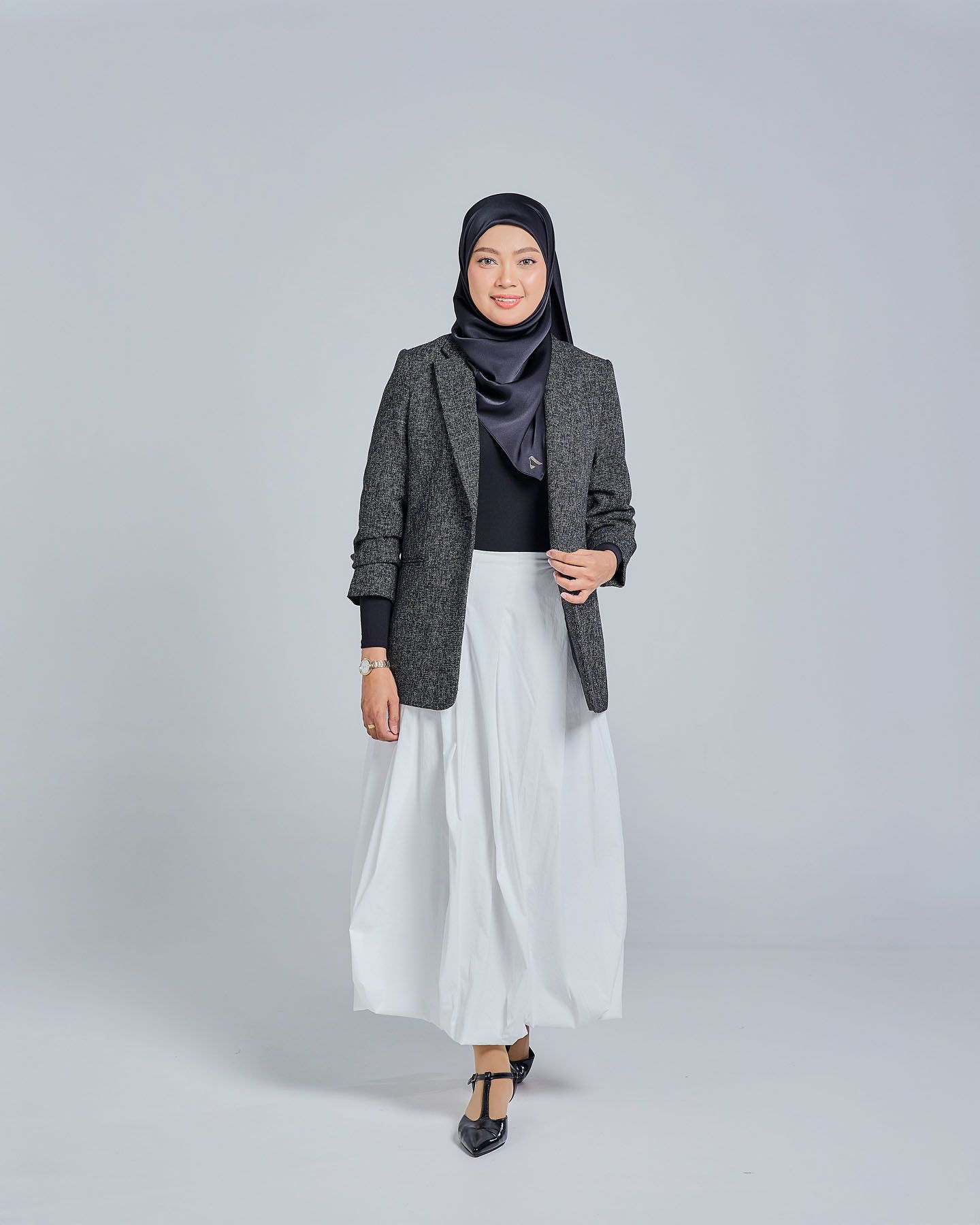 Muslim woman in gray herringbone blazer, black top, white maxi skirt, and navy hijab standing elegantly.