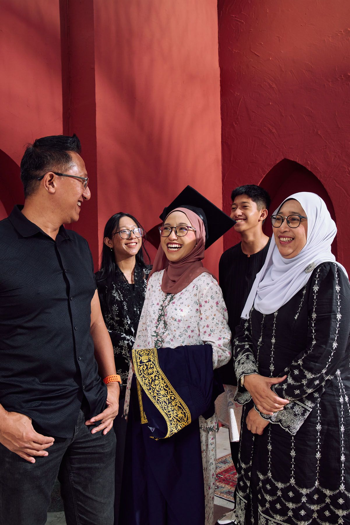 Muslim graduate in mortarboard laughs joyfully with family in red archway setting.