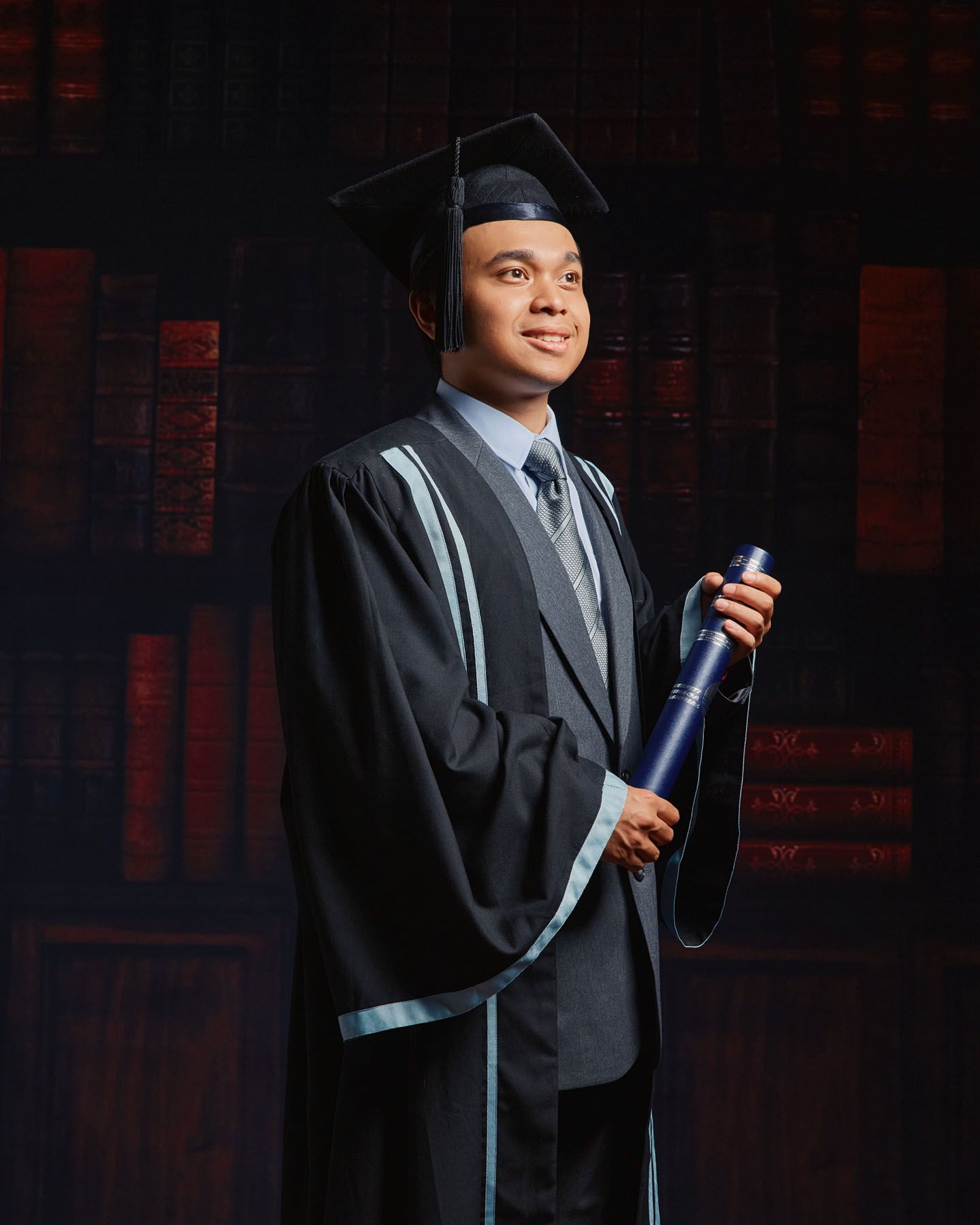 Male graduate in black academic gown and mortarboard holding diploma scroll in studio portrait.