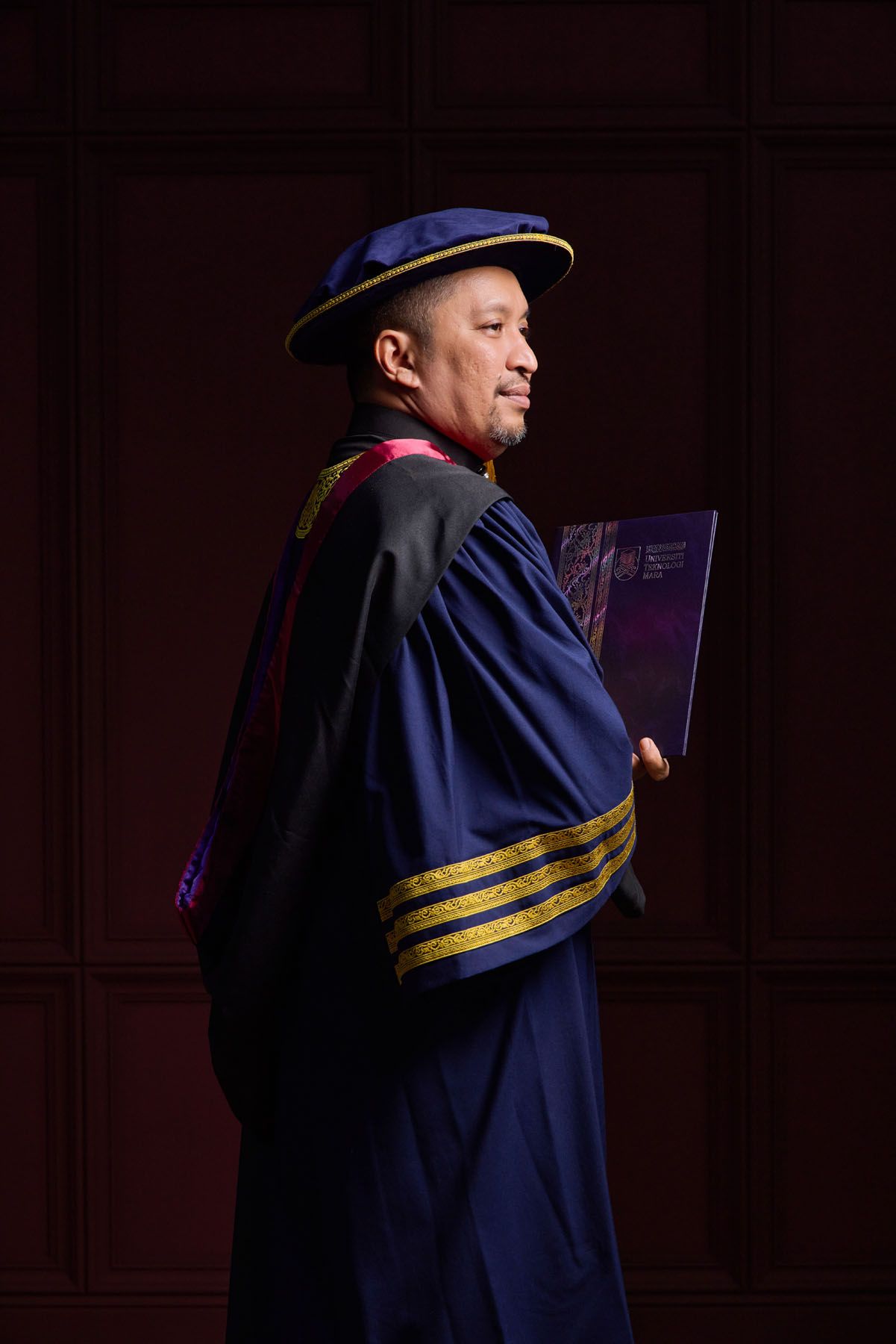 Man in blue doctoral graduation robe with gold stripes holding diploma book in side profile pose.