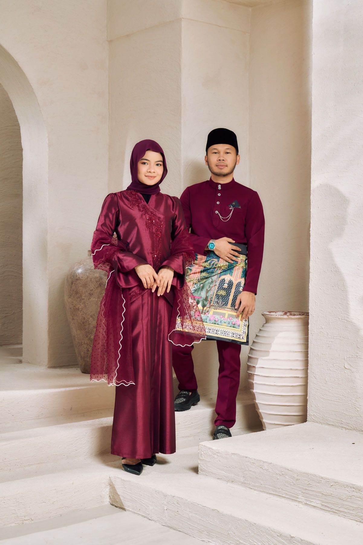 A Malaysian couple in matching burgundy traditional attire standing on steps beside decorative ceramic vases indoors.