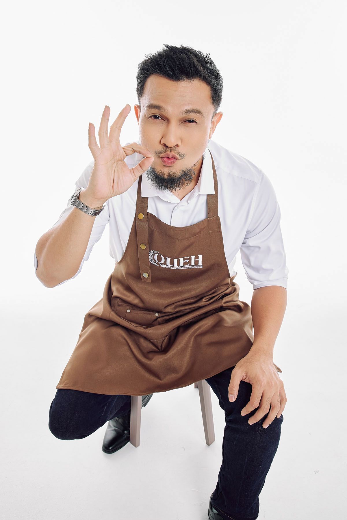Asian man in brown apron sitting on chair making OK gesture with kissing expression.