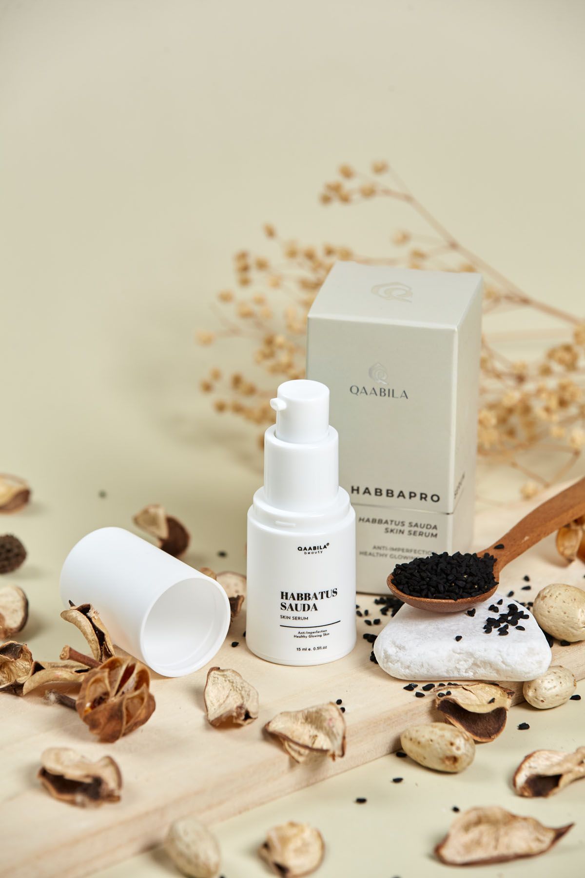 Qaabila Habbatus Sauda skin serum with black seeds on wooden spoon, dried botanicals on beige background.