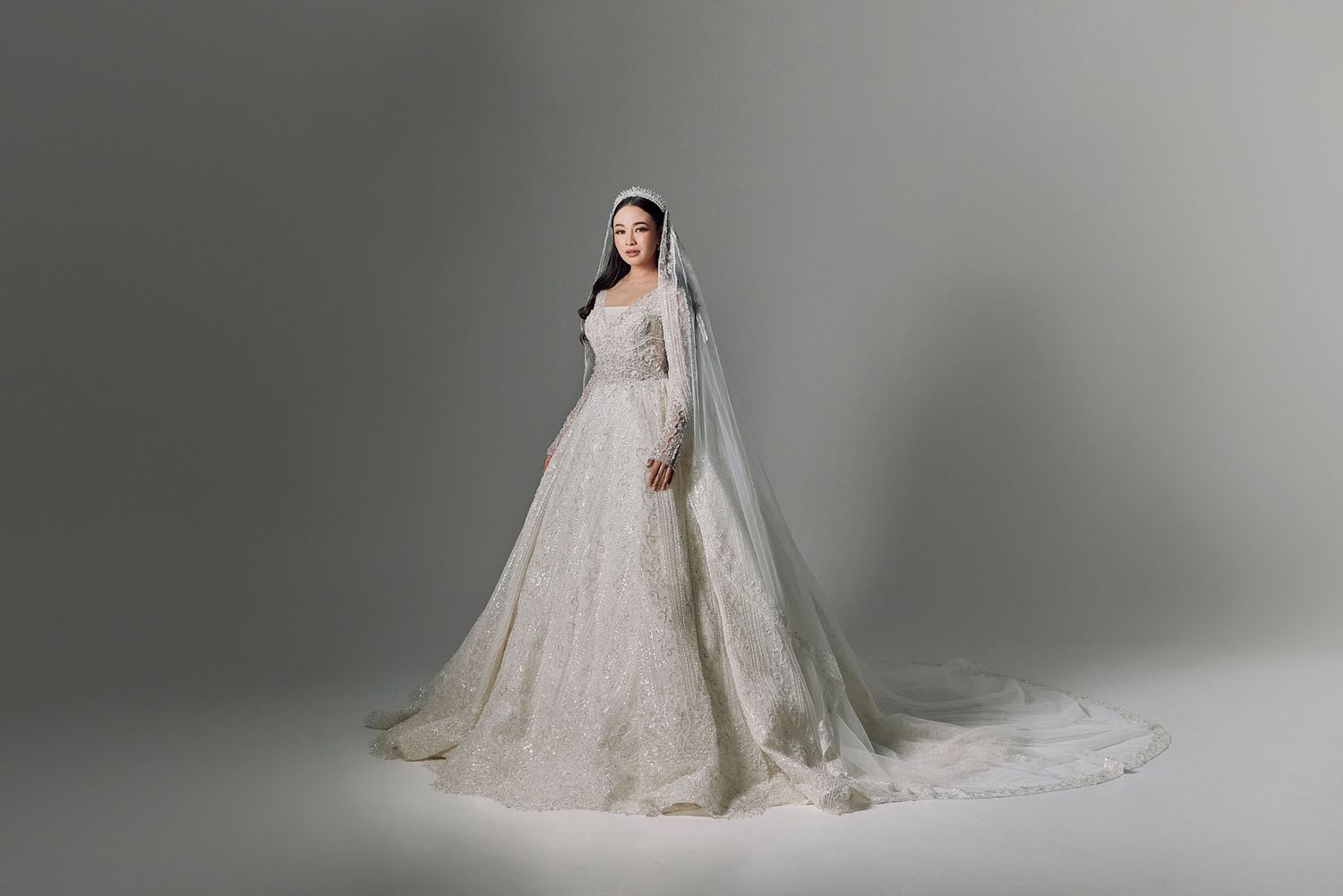 Muslim bride in a voluminous white lace ball gown with long veil posing gracefully on a grey background.